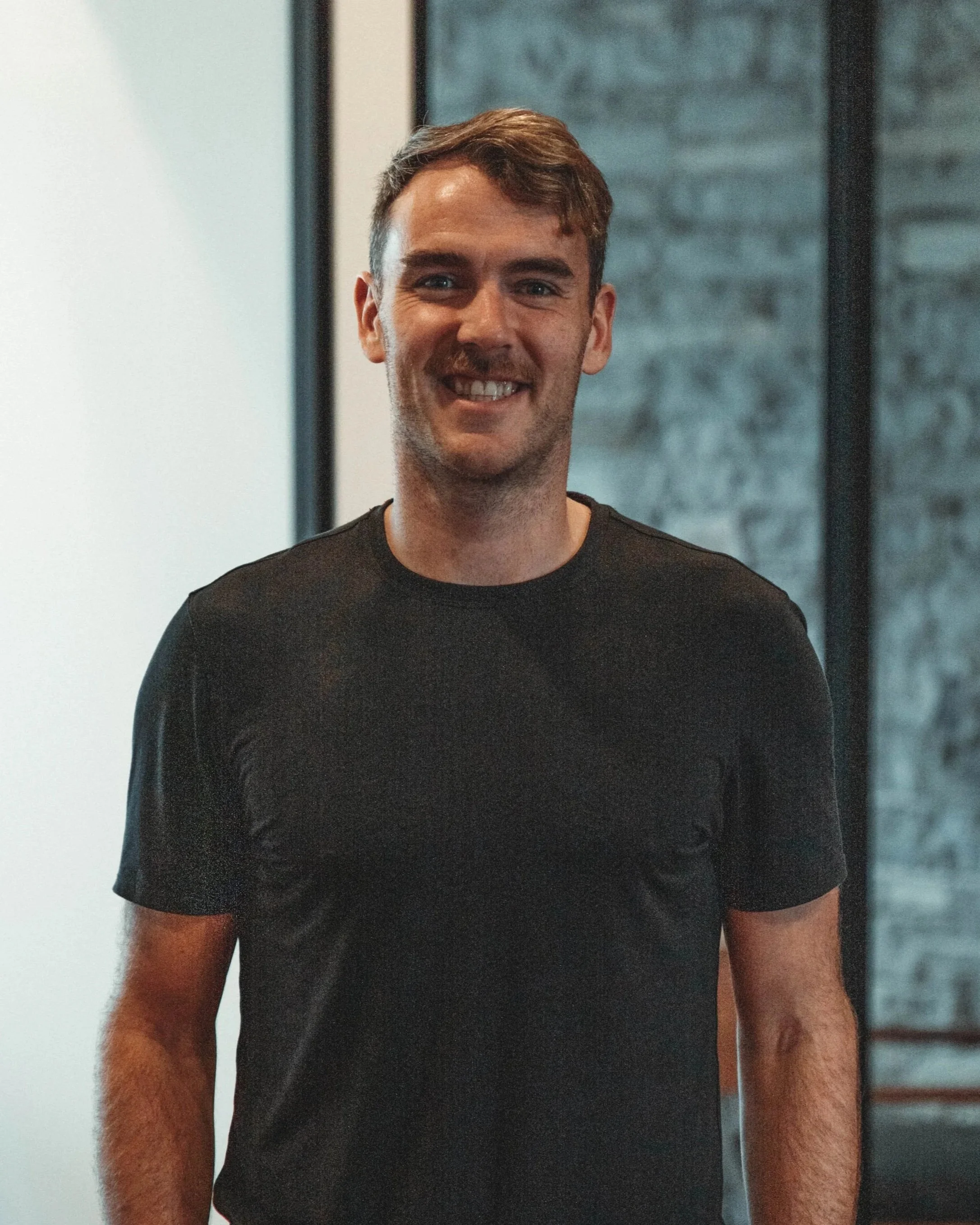 A young man with short dark hair, smiling, wearing a black t-shirt, standing in front of a brick wall.