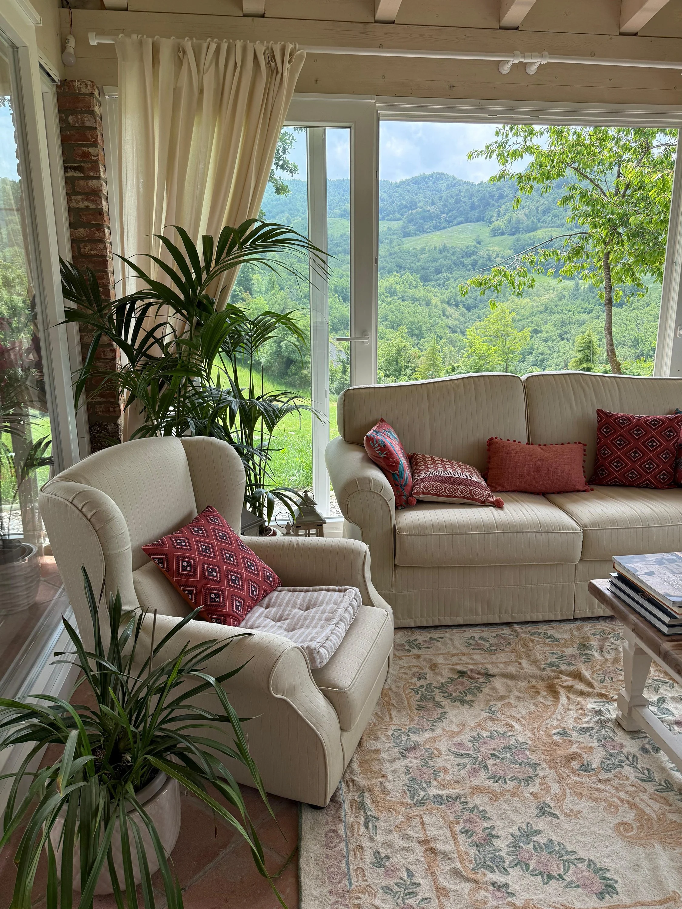 Interno di un soggiorno con divano bianco, cuscini rossi e bianchi, stufa a legna, tende beige, lampada da tavolo su un mobile rosso e finestre panoramiche con vista sul verde esterno.