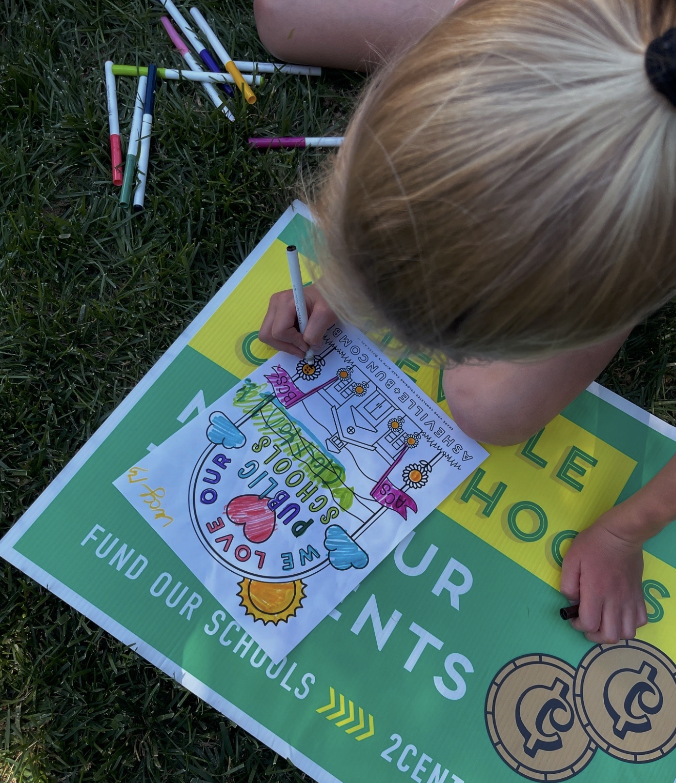 A child with blonde hair drawing on a colorful flyer outdoors on grass, surrounded by markers. The flyer promotes supporting schools and includes drawings of hearts and sunflowers.