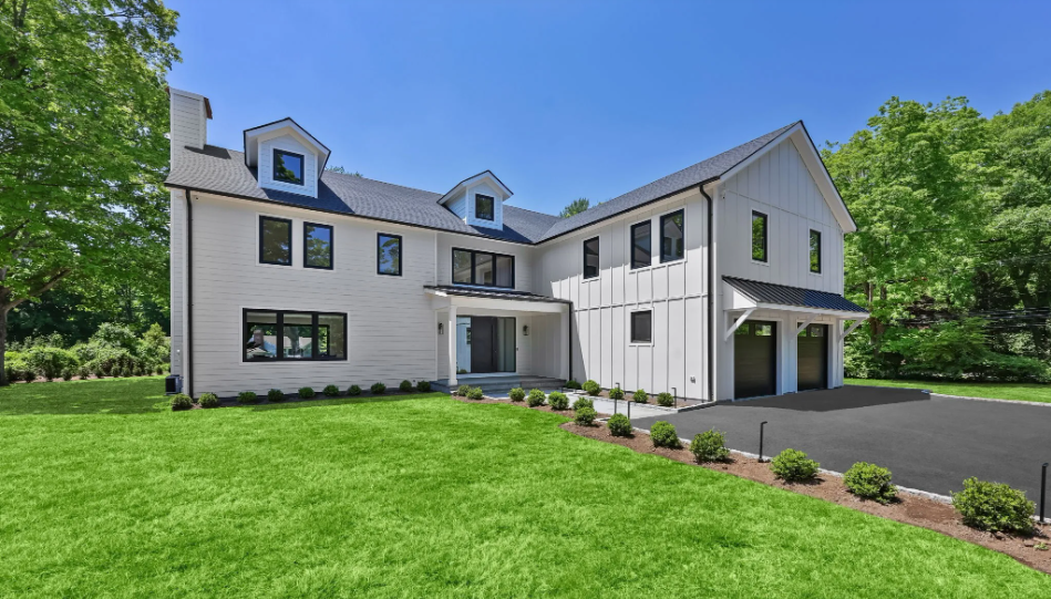 Modern two-story house with white exterior, black roof, multiple windows, attached garage, surrounded by green lawn and trees under a clear blue sky.