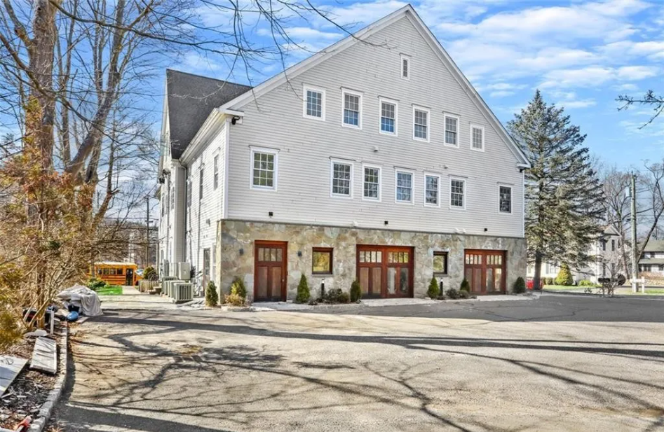 Large white building with stonework on the lower level, multiple windows, and a sloped roof, surrounded by trees and an asphalt parking area.