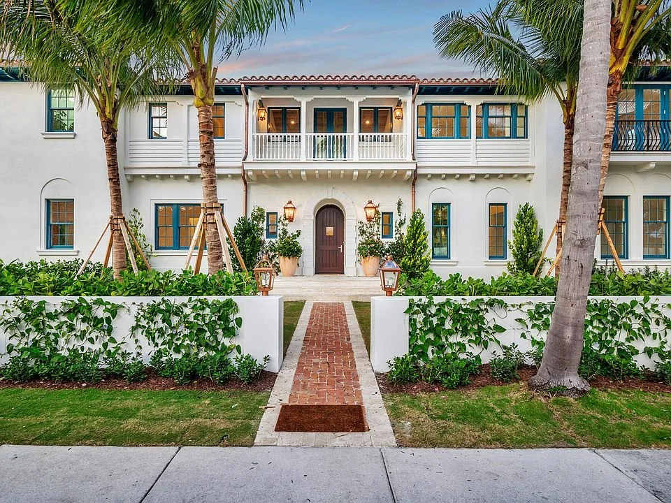 Front view of a white, two-story Mediterranean-style house with palm trees, lush greenery, and a brick pathway leading to the main door.