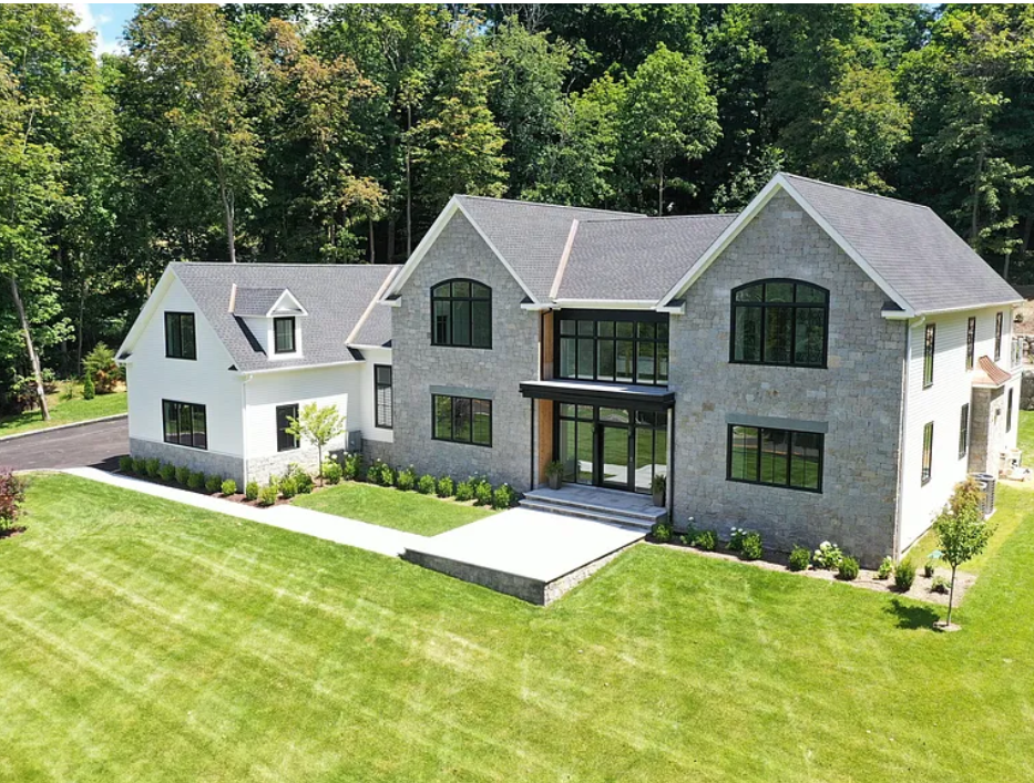 A modern two-story house with a stone facade, large windows, a front porch with stairs, and well-maintained lawn surrounded by trees.