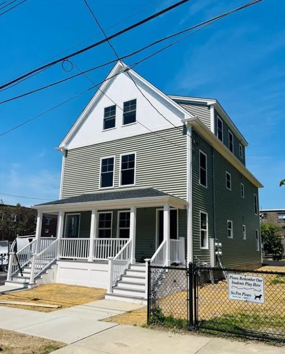 A three-story house with a white upper section and gray lower section, featuring a front porch with white railings and stairs, black-framed windows, and overhead power lines against a blue sky.