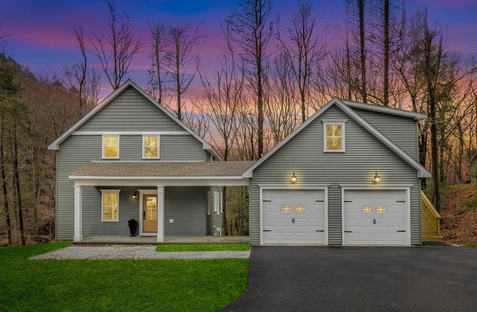 A two-story gray house with a front porch, attached two-car garage, and a driveway, set against a backdrop of trees during sunset.