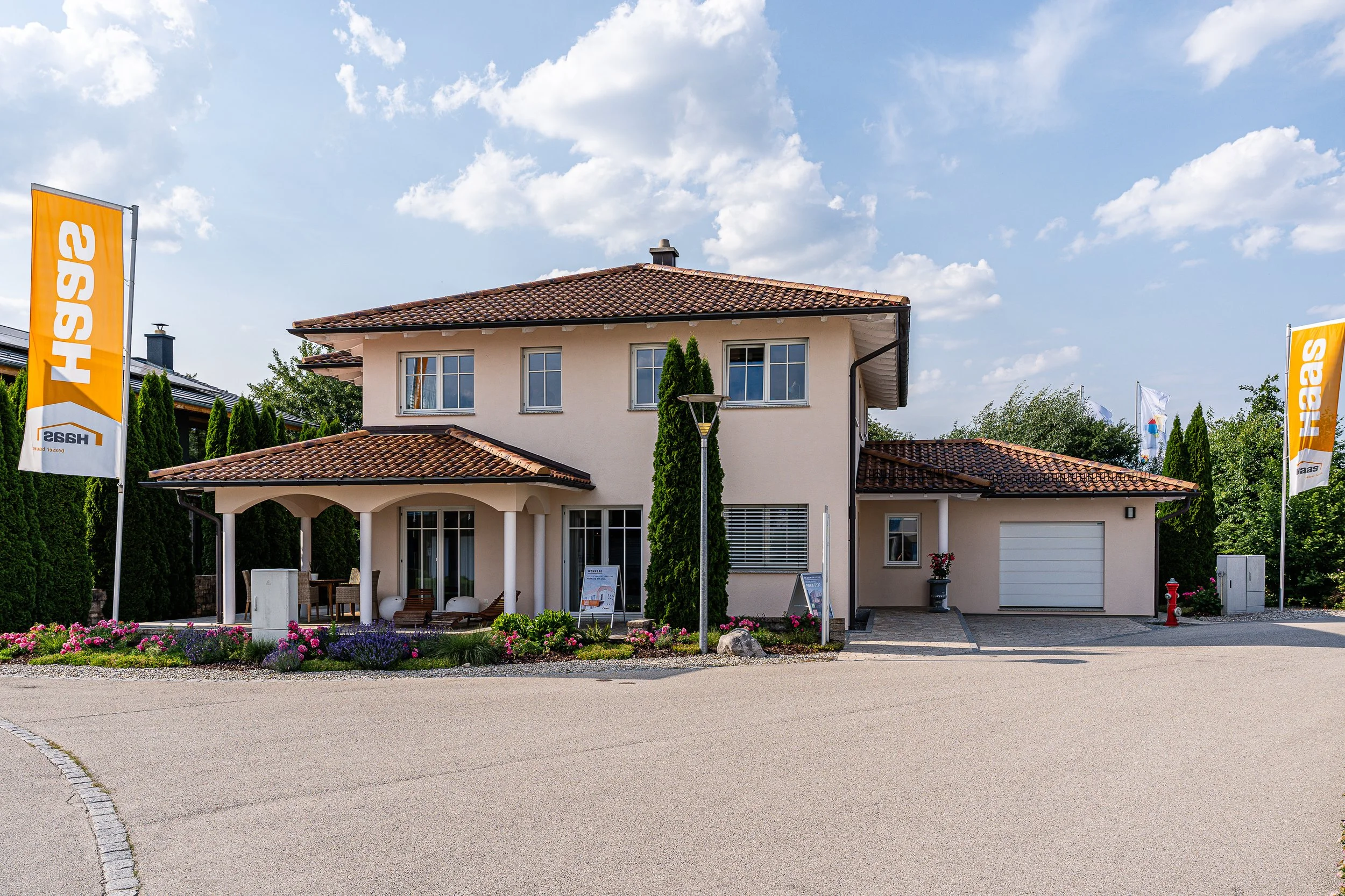 Haas Musterhaus Poing im Toskana Stil. Zwei Fahnen mit 'Haas' vor einem modernen, zweistöckigen Haus mit rotem Ziegeldach und türkisen Fensterrahmen, umgeben von Garten mit Blumen, auf einer gepflasterten Straße bei sonnigem Himmel.