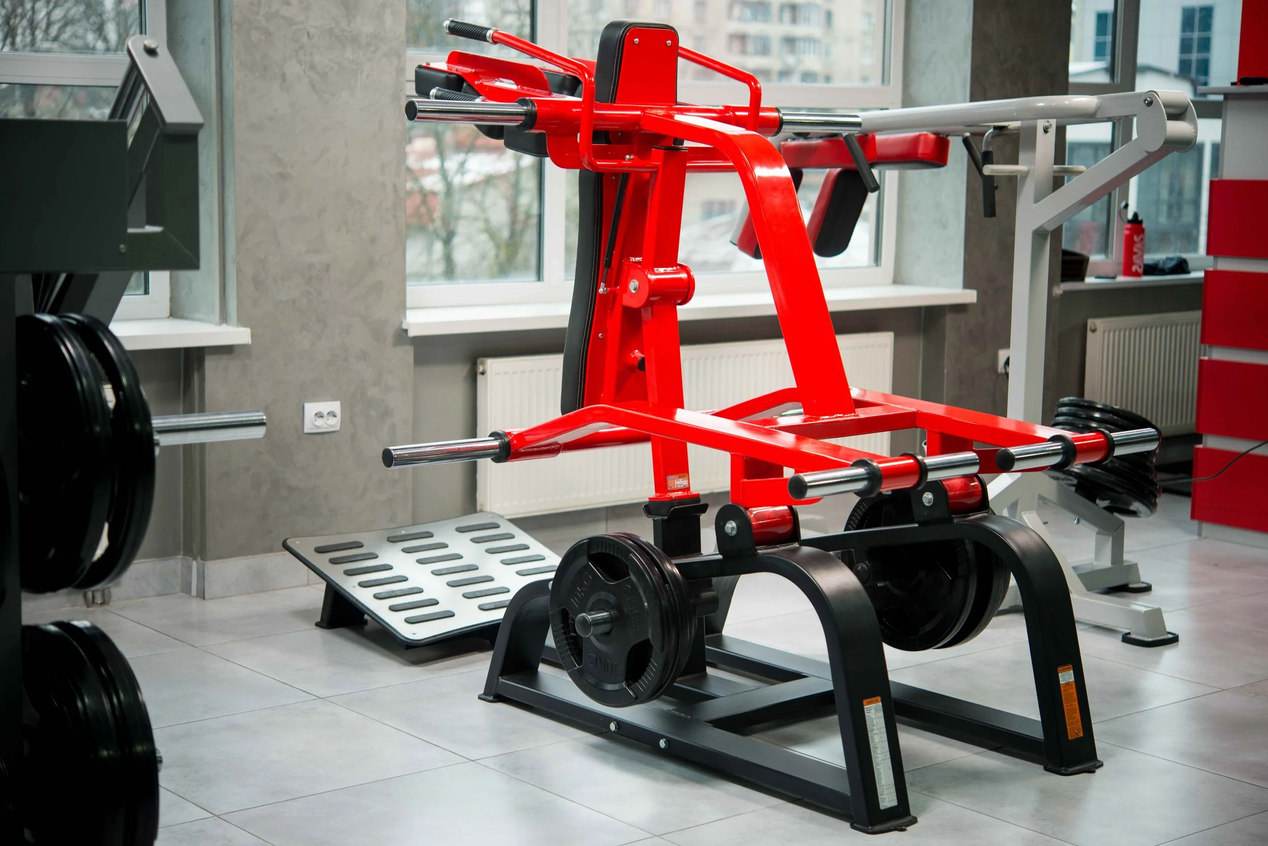 Red and black weightlifting resistance equipment in a gym with large windows and a gray concrete wall.