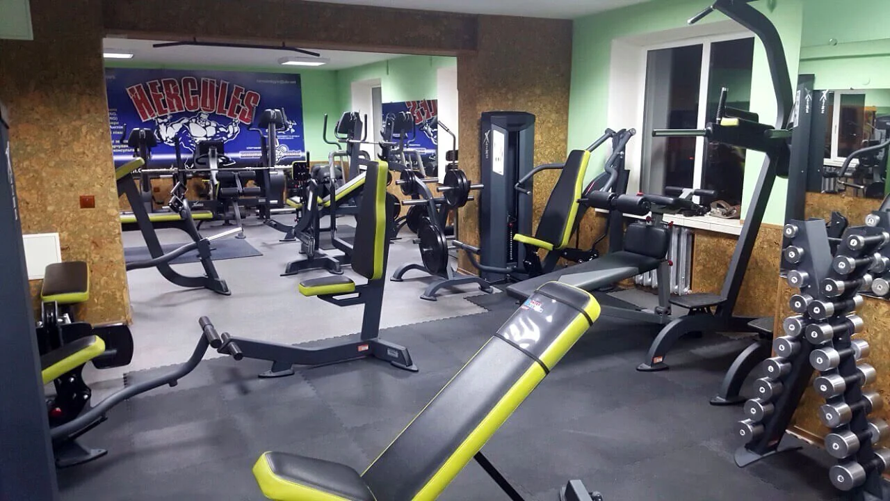 Empty gym with various workout equipment including weight benches, a rack of dumbbells, and cardio machines, with mirrors and a Hercules sign on the wall.