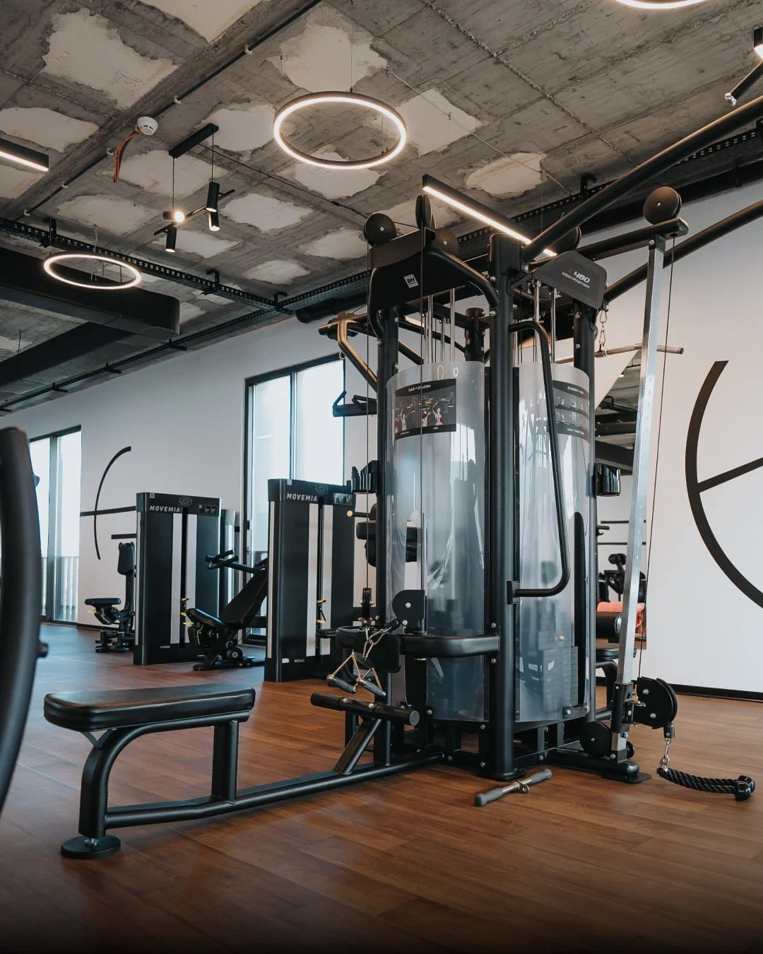Modern gym with workout equipment, wood flooring, exposed ceiling with circular and linear lighting, and large windows.