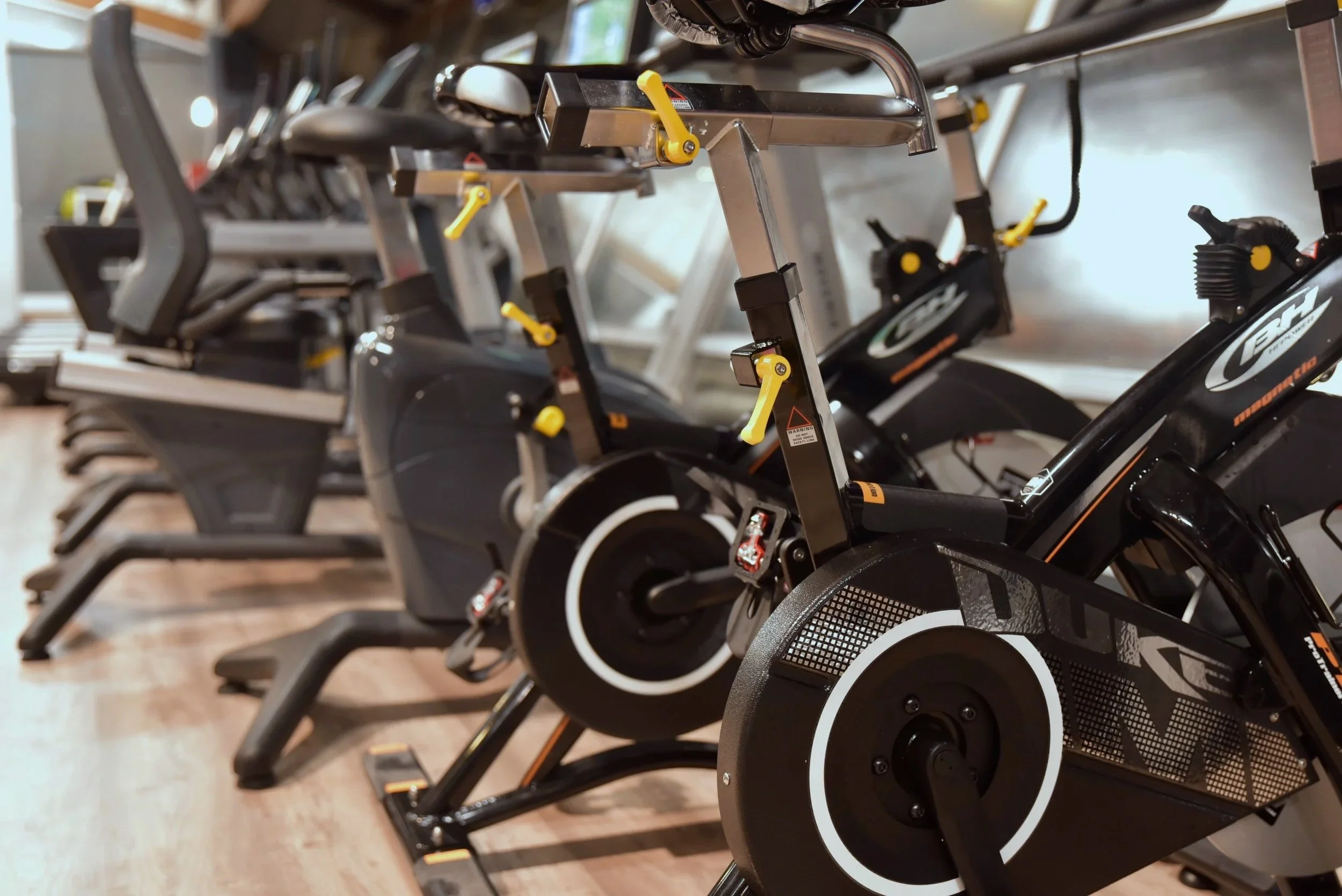 Indoor gym with multiple stationary bikes aligned in a row, focusing on the black and white fitness bikes with yellow adjustment levers.