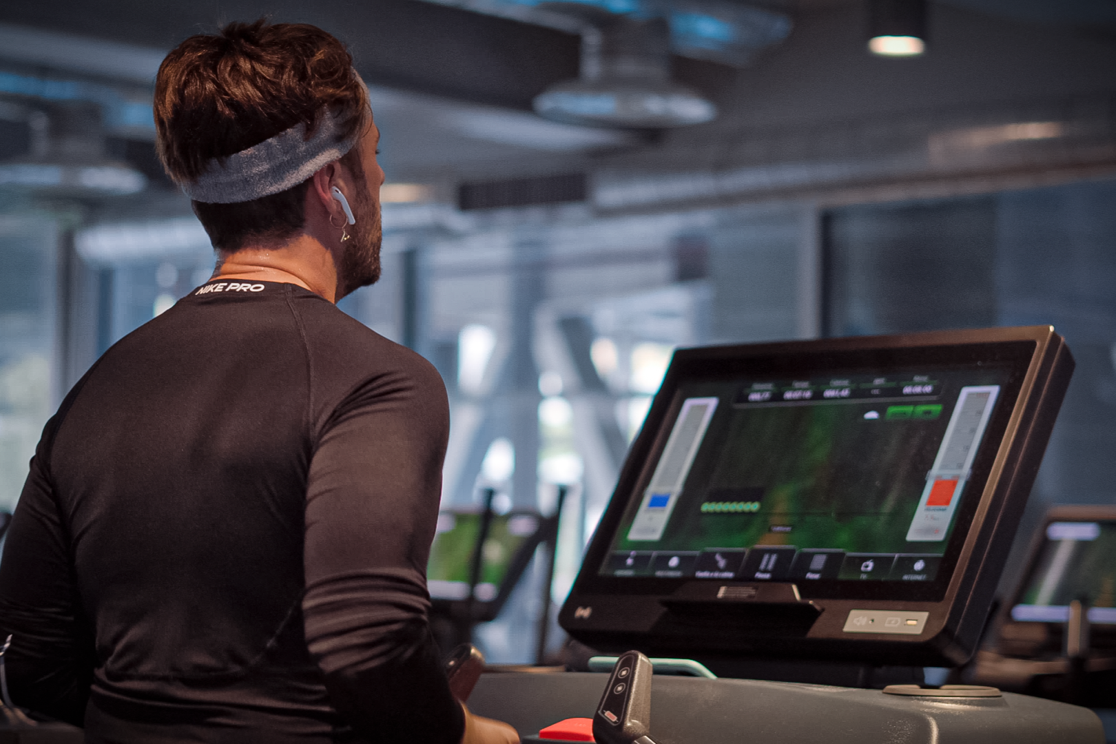 A man with brown hair, beard, wearing a headband, wireless earbuds, and a black long-sleeve shirt, is standing at a treadmill with a digital screen in an indoor gym.