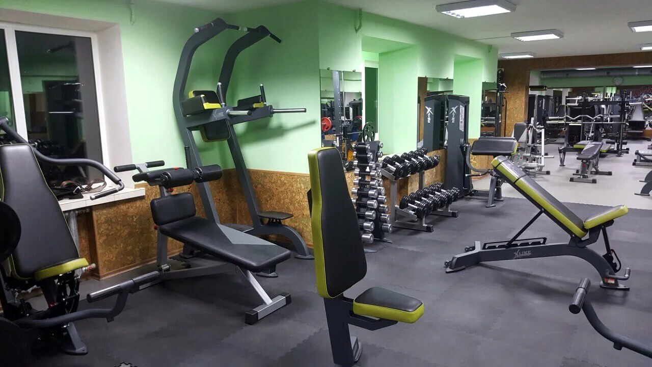 Empty gym with various workout equipment including weight benches, dumbbell racks, and exercise machines, in a room with green walls.