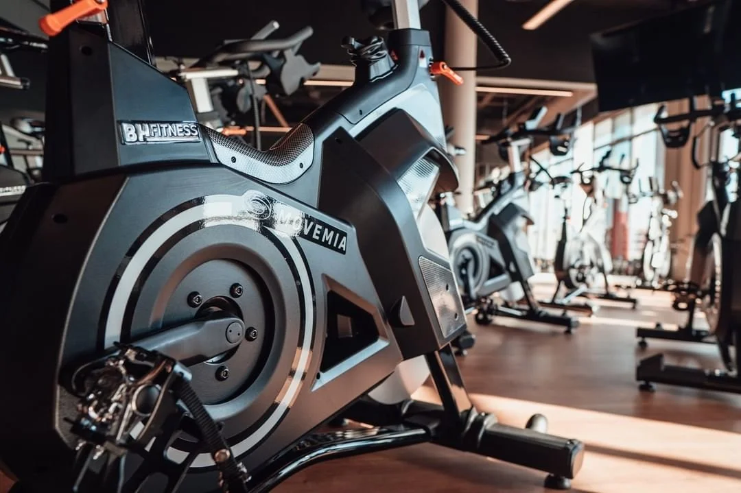Close-up of an exercise bike in a gym with more fitness equipment in the background.