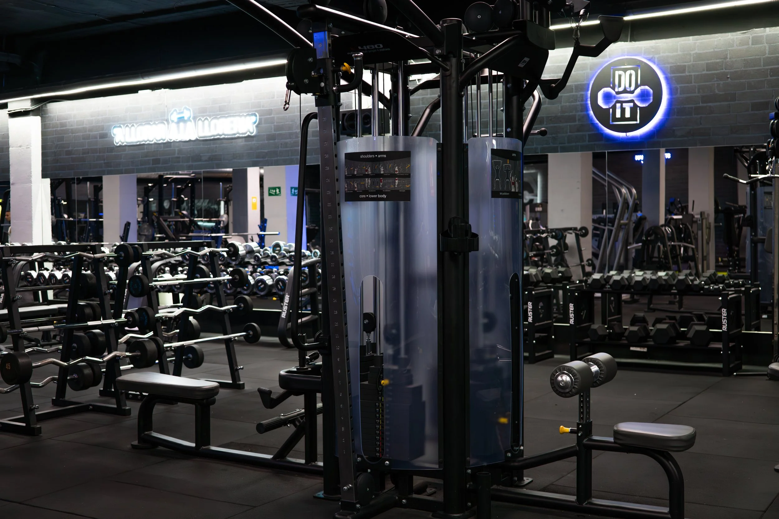 A gym with various workout equipment, including dumbbells, benches, and a multi-function weight machine, with neon signs on the wall that say 'Welcome to the Gym' and 'Do It'.