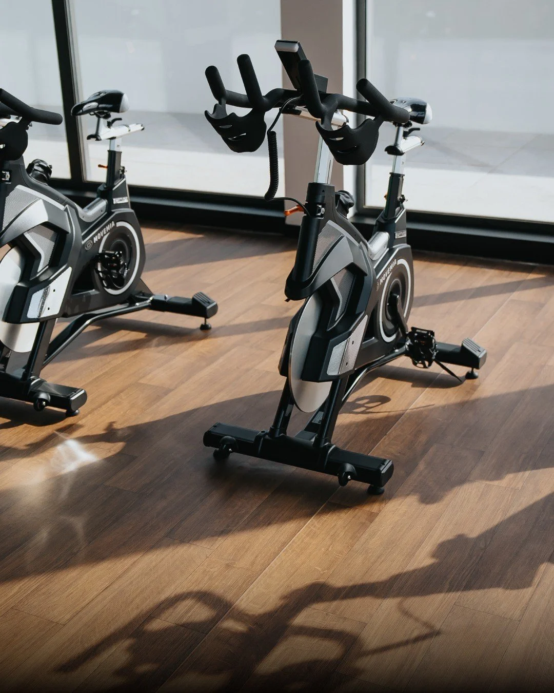 Two stationary exercise bikes in a brightly lit room with wooden flooring and large windows.