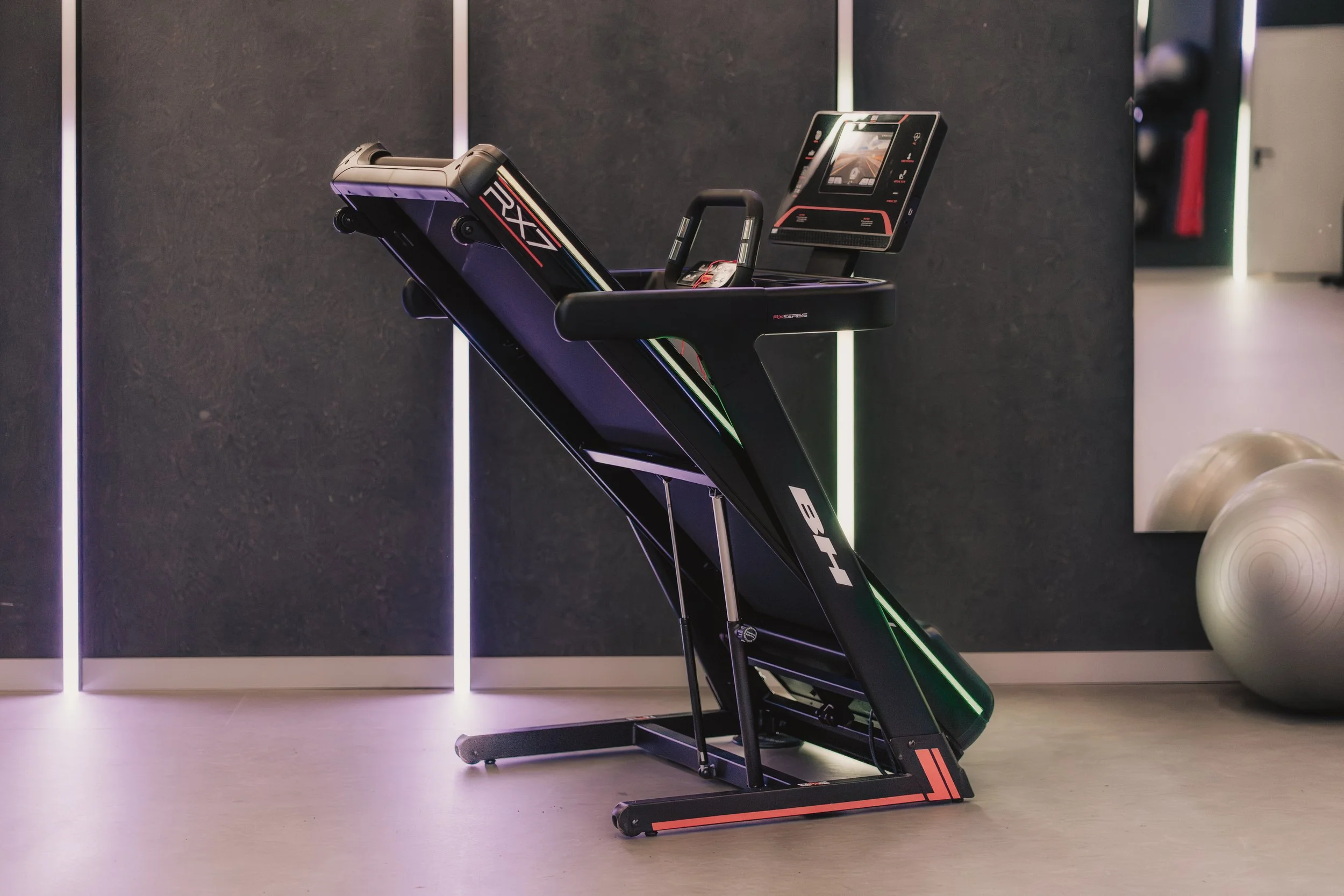 A modern treadmill with a digital display in a gym, illuminated by colorful lights and workout balls in the background.