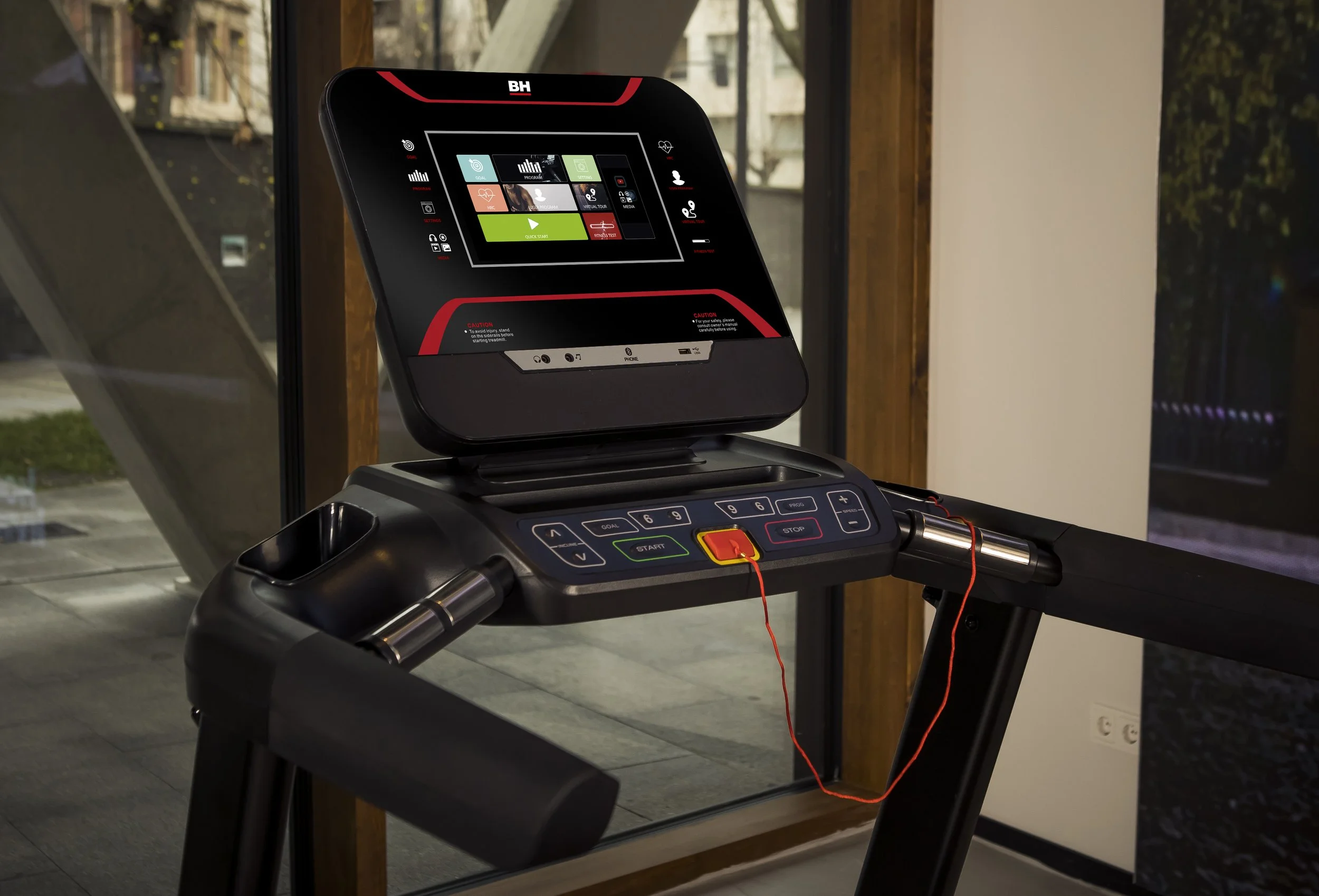 Close-up of a treadmill's control panel and screen, with red safety key attached by a cord, situated near a window with a view of the outdoors.
