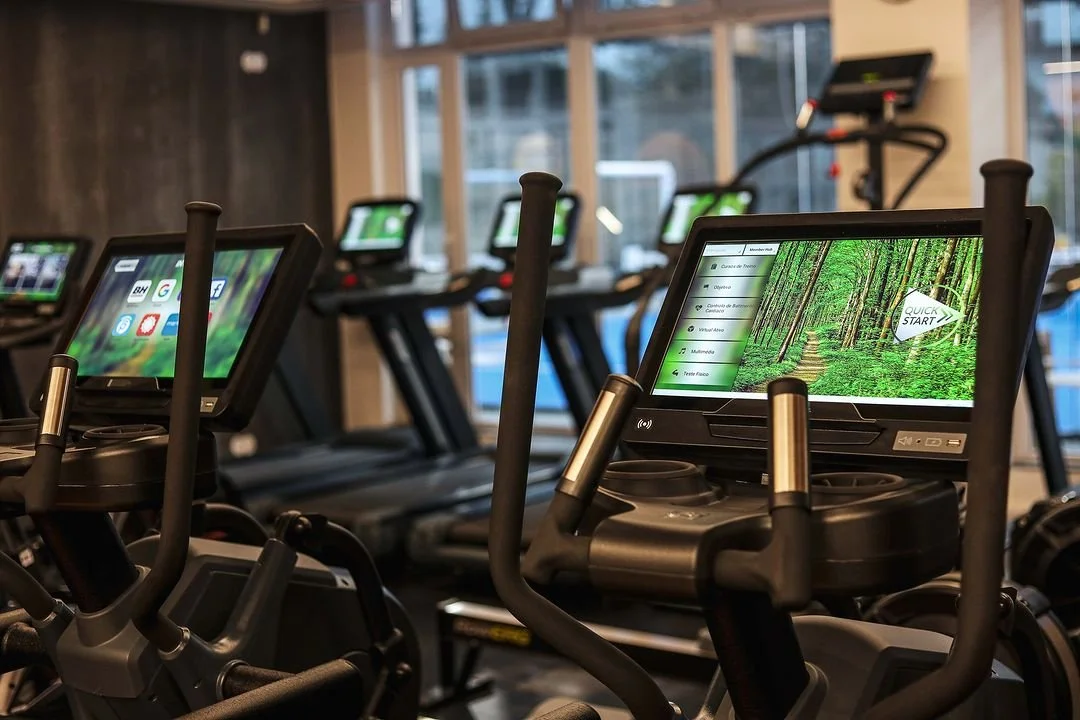 Row of modern treadmills in a gym, each equipped with a digital screen, with a view of large windows in the background.