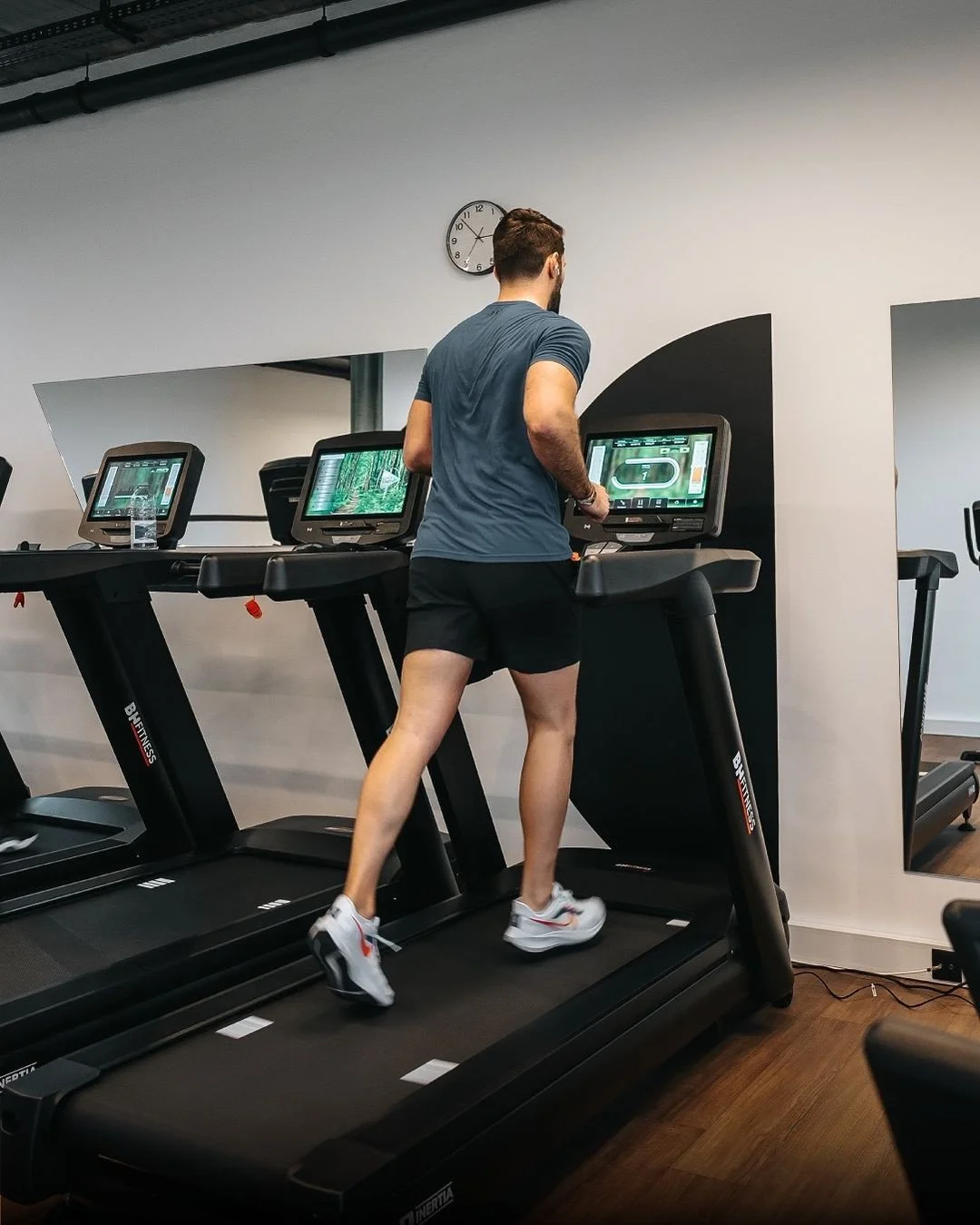 A man running on a treadmill in a gym, surrounded by multiple treadmill screens, with a white wall, black accents, and a clock on the wall.