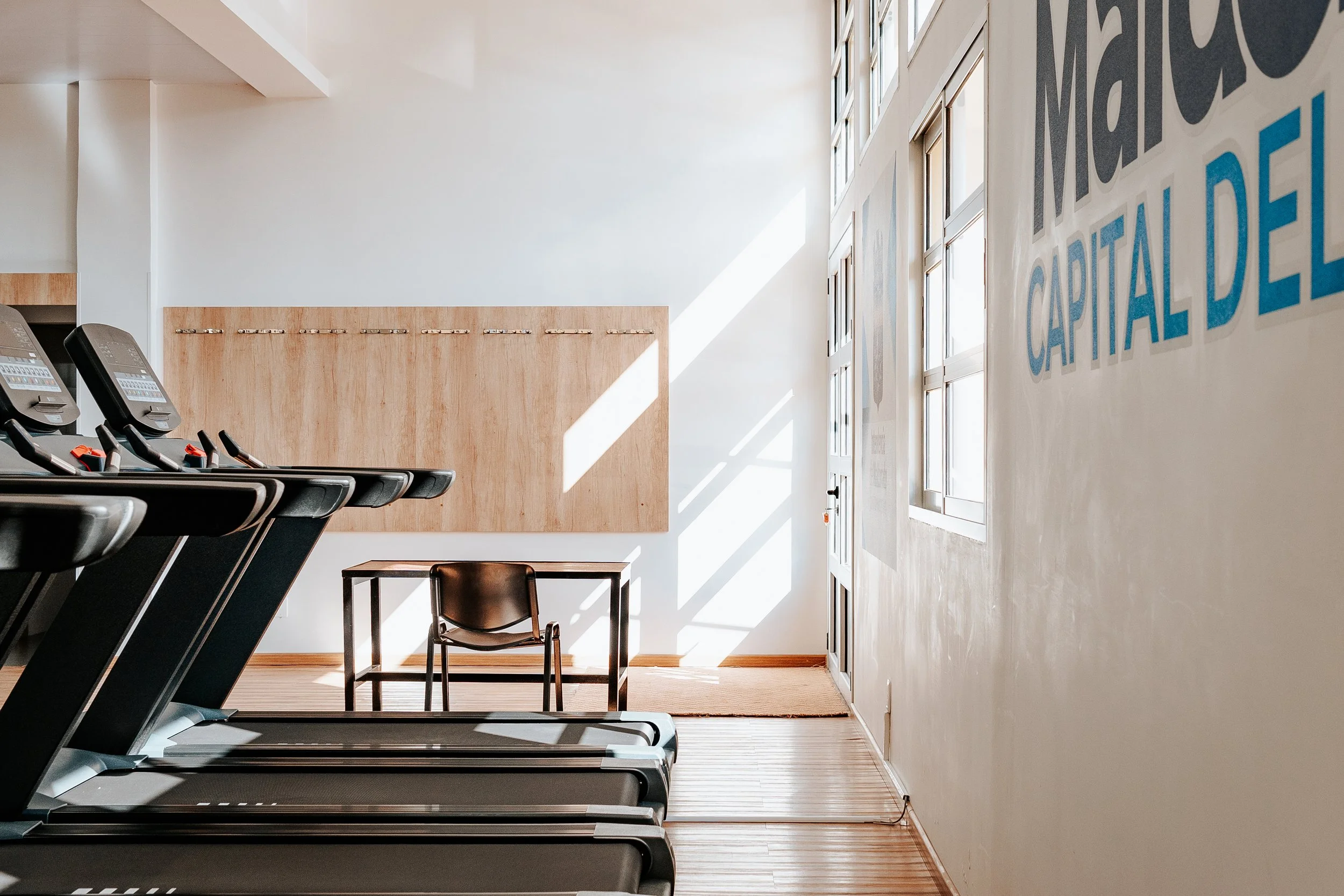 A fitness gym with treadmills on the left and a small office desk and chair near the back wall, illuminated by sunlight through windows on the right.
