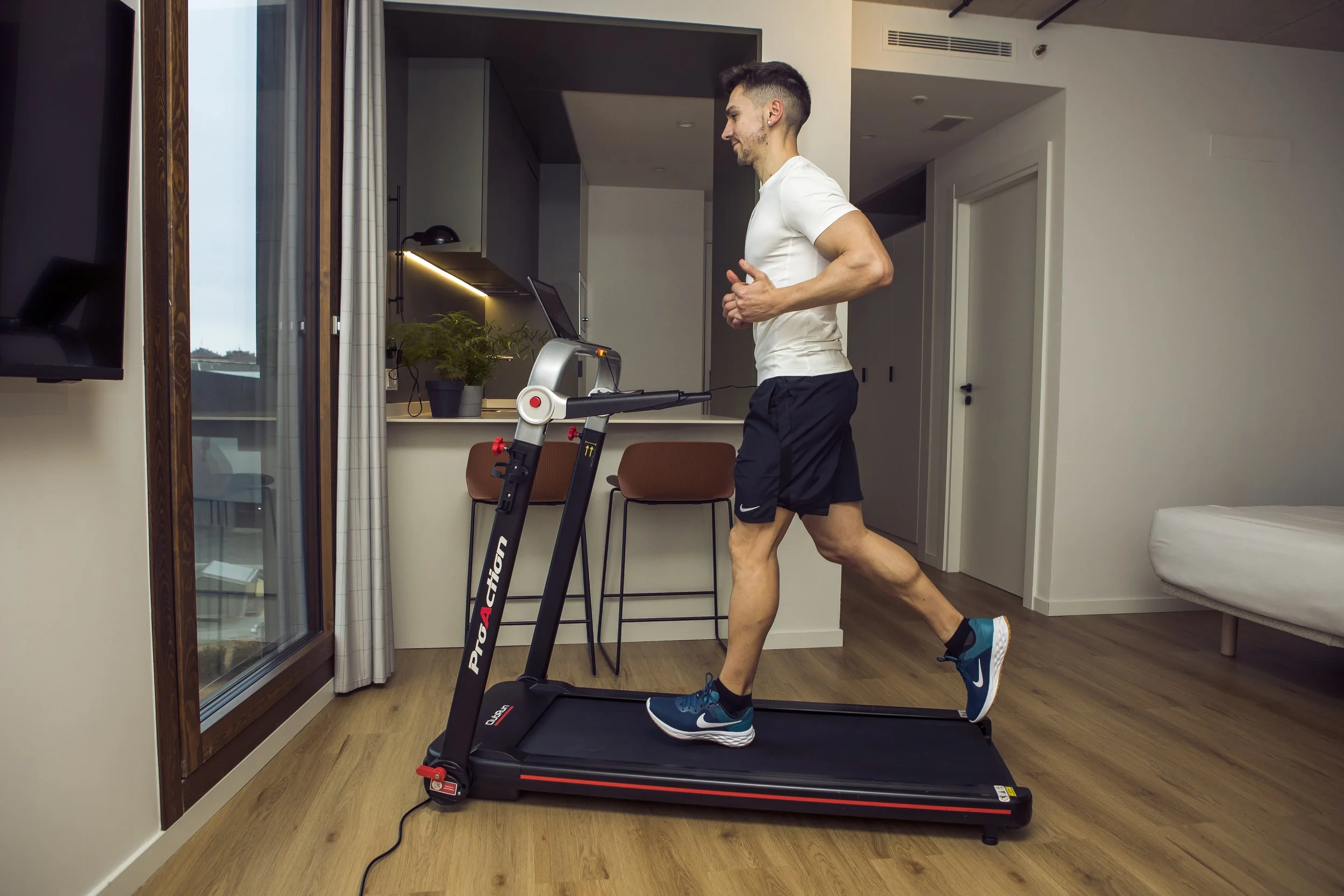 Man running on a treadmill inside a modern apartment with wooden floors, a sliding glass door, and minimal decor.