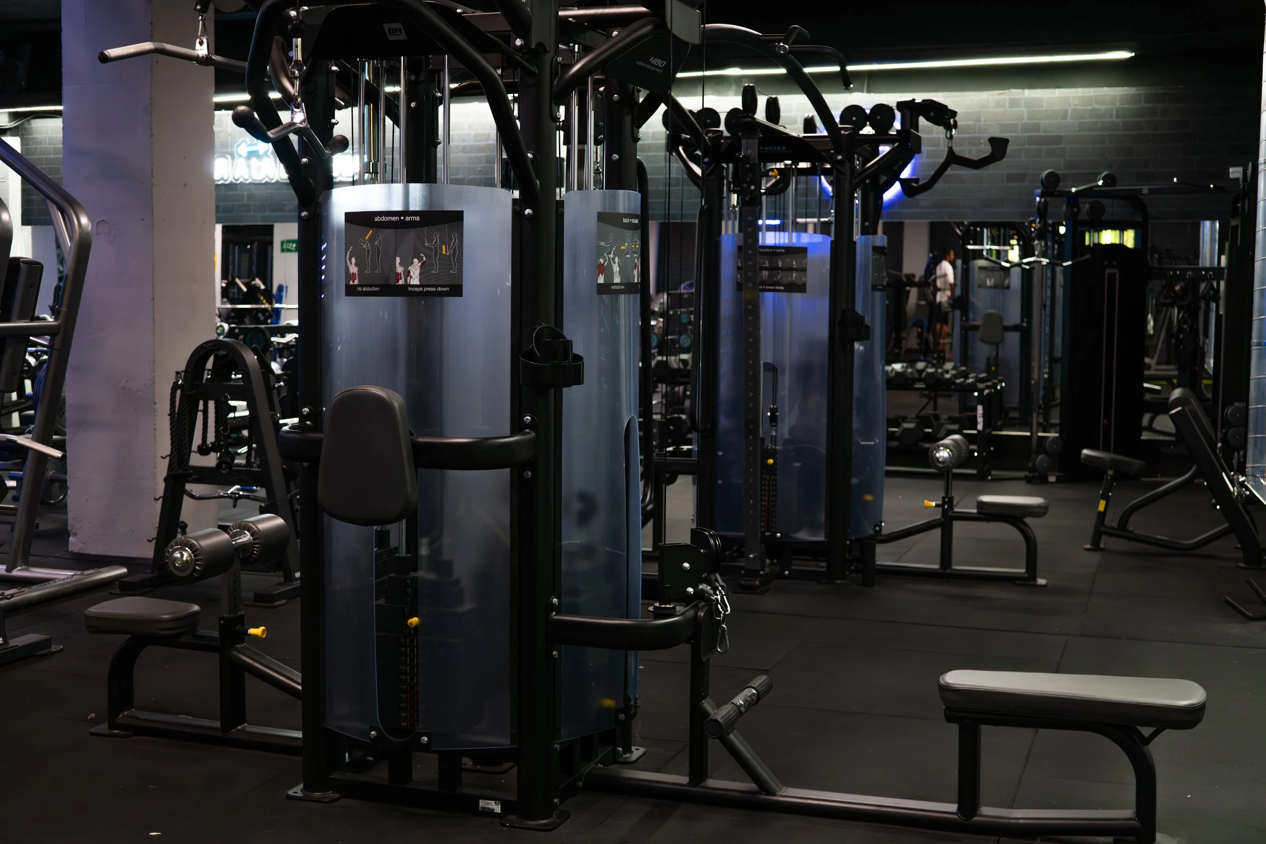 Gym with various workout equipment including weight machines, benches, and a person in the background. Brick wall and neon signs are visible.