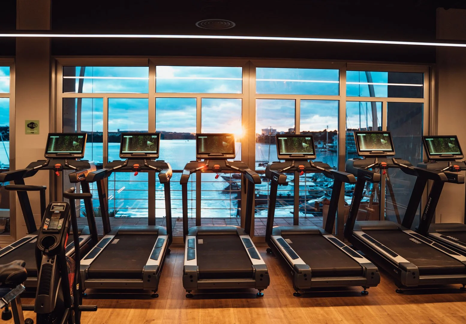 Indoor gym with five treadmills facing large windows overlooking a marina with boats at sunset.