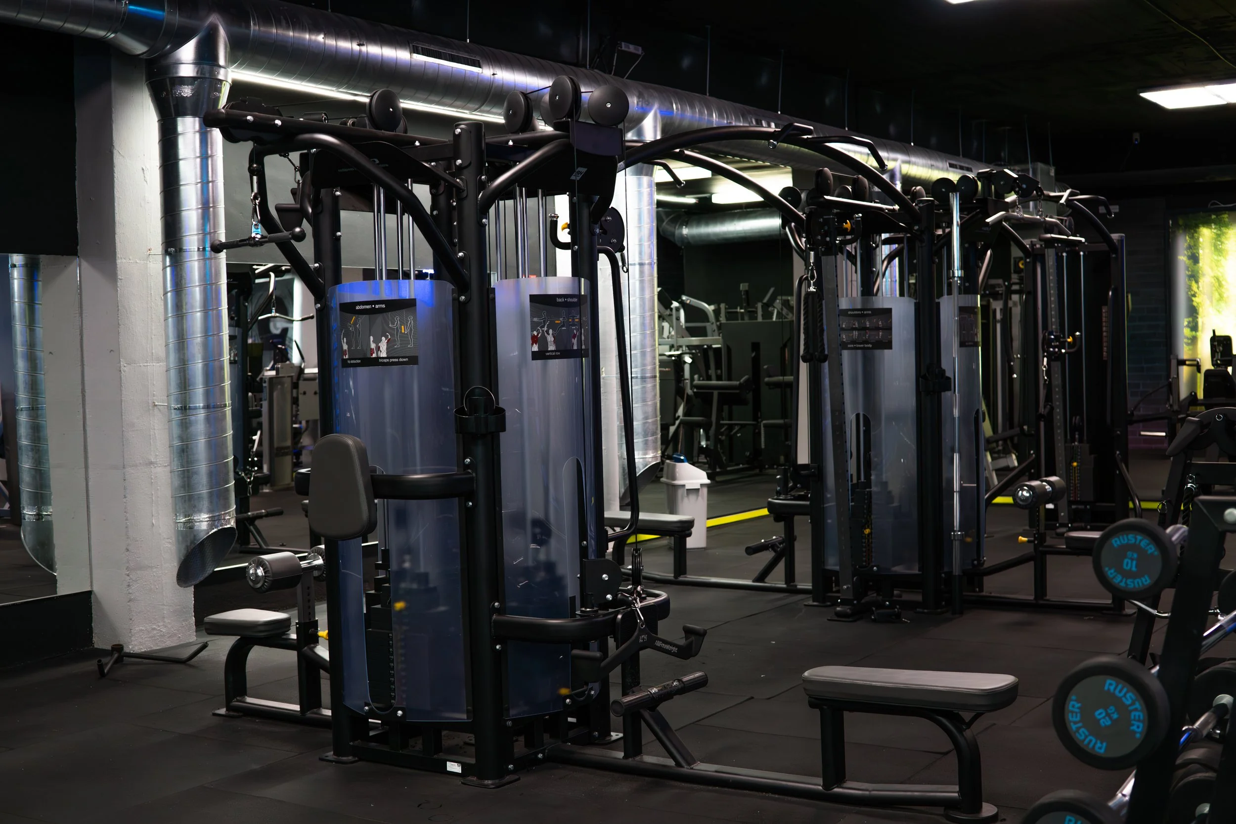 Empty gym with various workout equipment including weight machines and free weights, with black flooring and industrial style pipes on the ceiling.