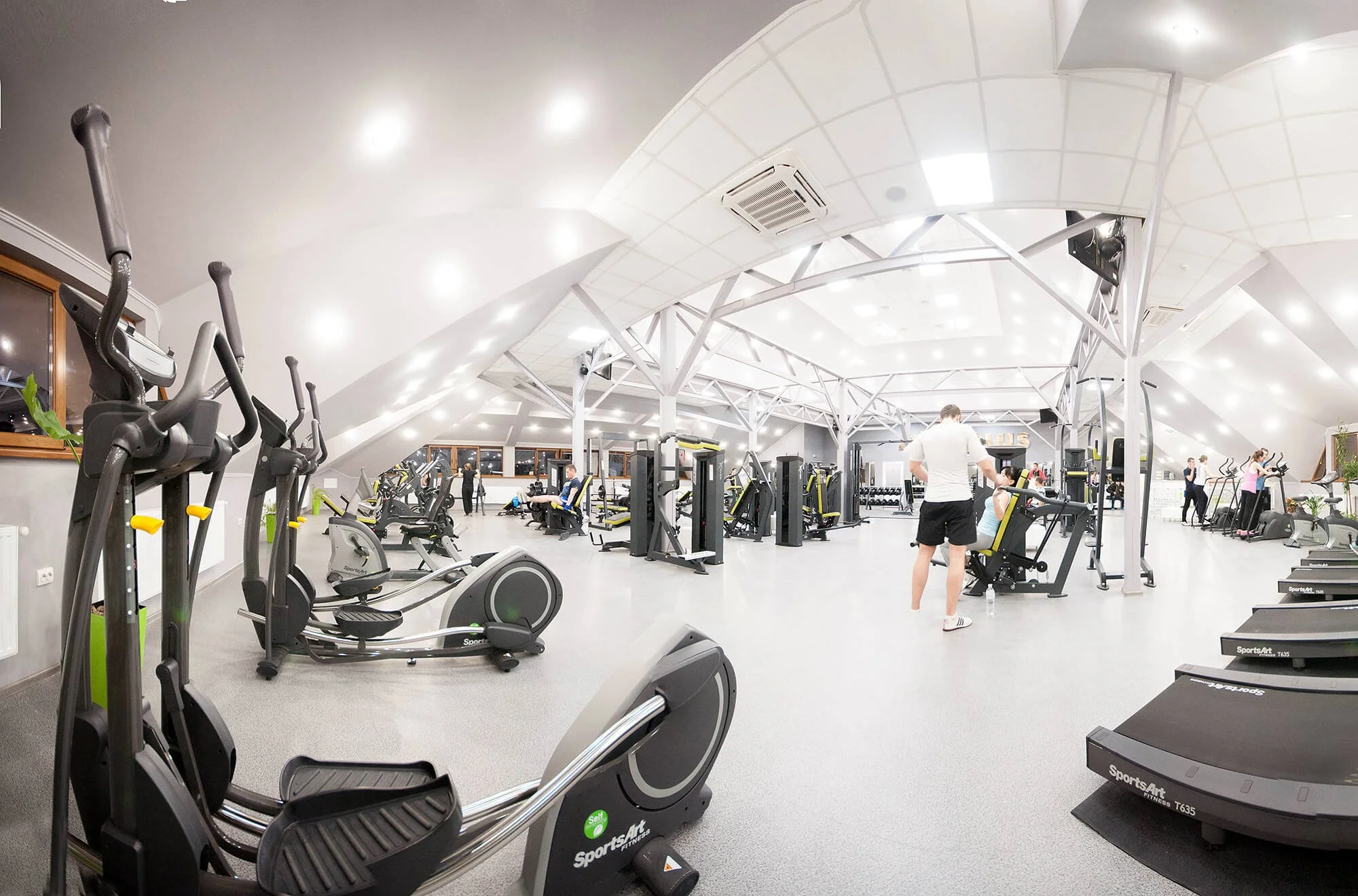 Interior of a modern gym with various exercise equipment, including treadmills, elliptical machines, and weight machines, with people working out and a bright, well-lit space with a white ceiling and large windows.