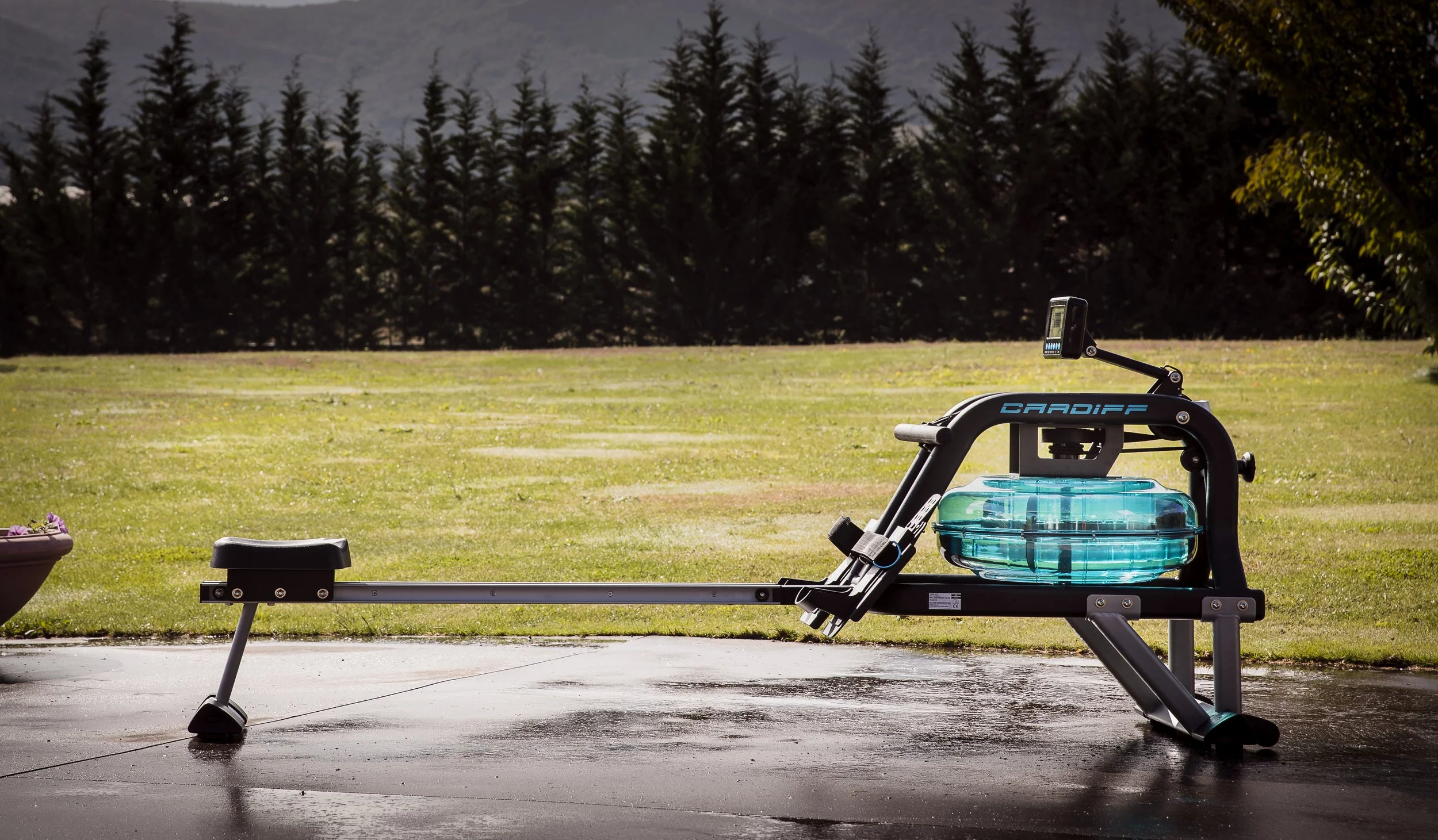 An outdoor ice water rowing machine on a wet black surface with grass and trees in the background.
