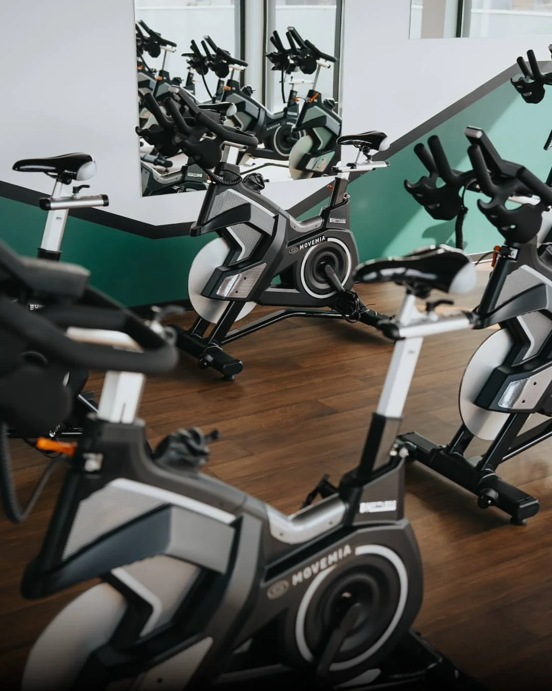 Indoor cycling studio with multiple stationary exercise bikes facing a mirror, with a wooden floor and a green and white wall.