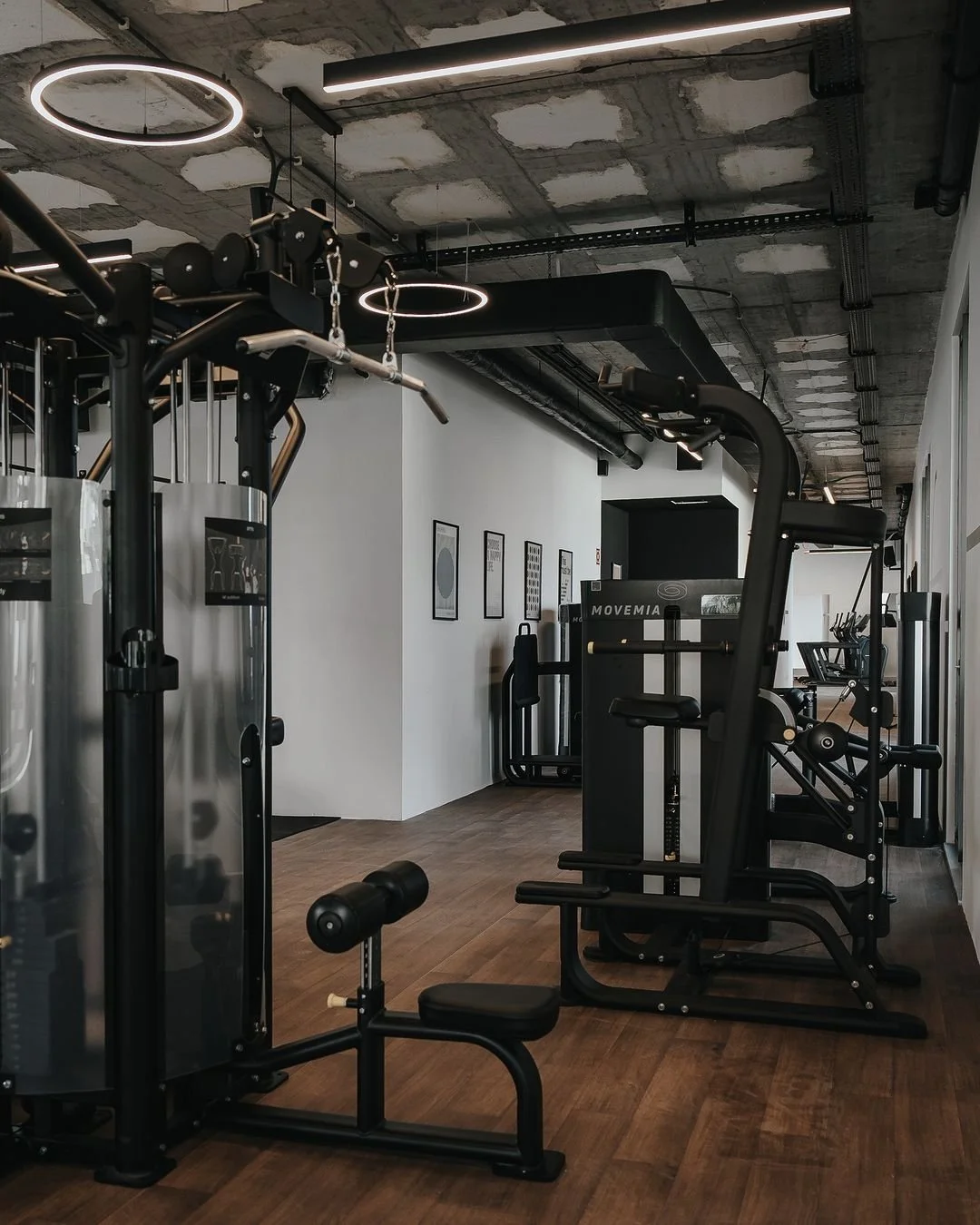 Interior of modern gym with various exercise machines on wooden floor, white walls, and exposed concrete ceiling.