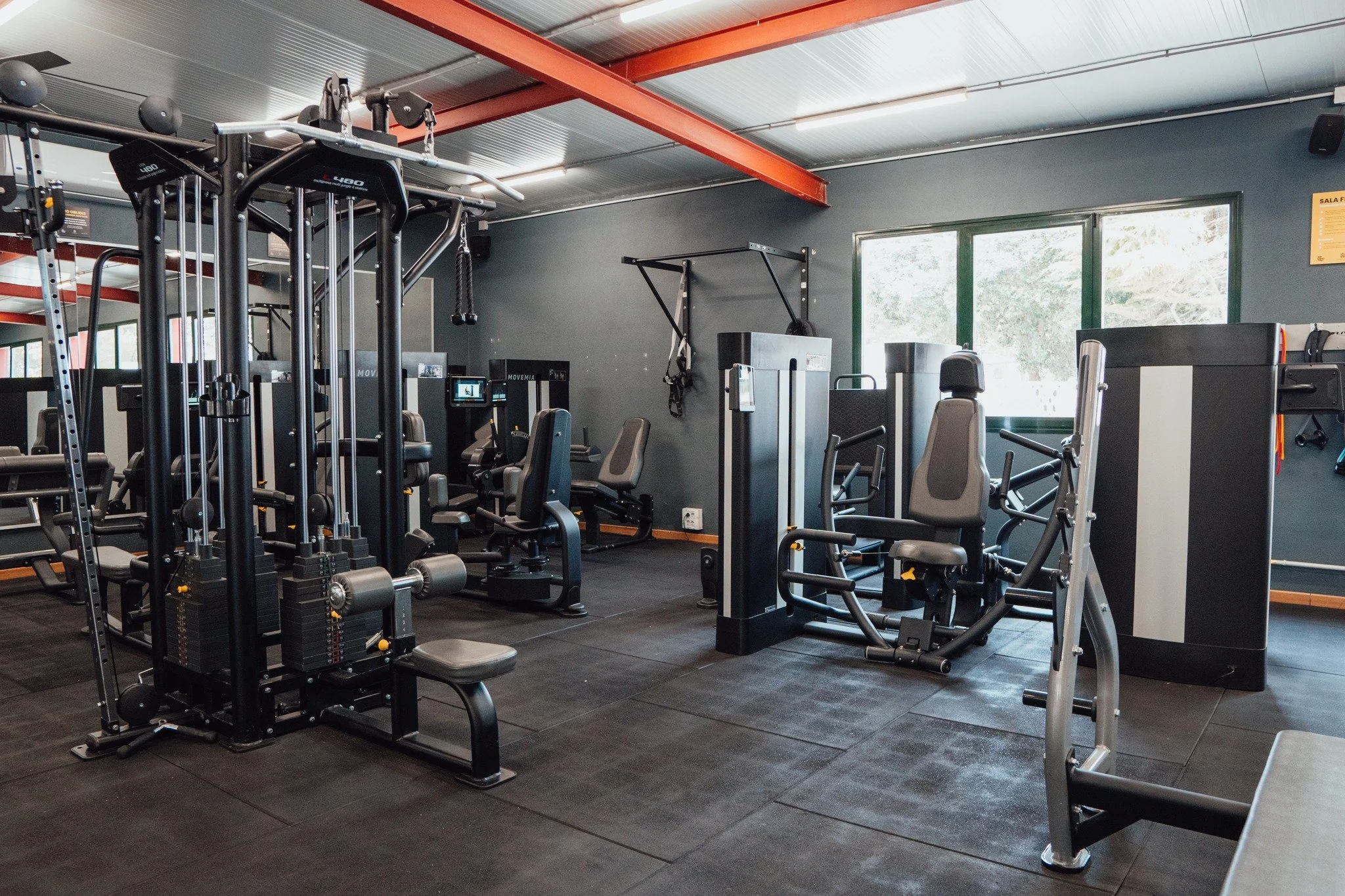 Empty gym with various exercise machines including weight training equipment and cardio machines, with black flooring and gray walls, large windows letting in natural light.