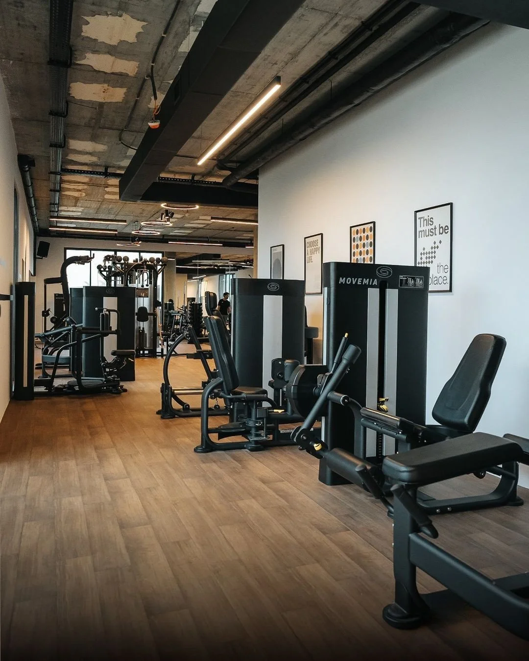 Fitness gym with various exercise machines arranged along a white wall with framed motivational posters, wooden flooring, exposed concrete ceiling, and a person working out in the background.