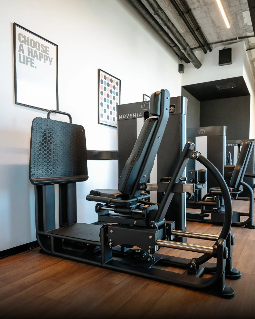 Indoor gym with black workout equipment, framed motivational posters on the white wall, concrete ceiling with exposed pipes, and hardwood floors.