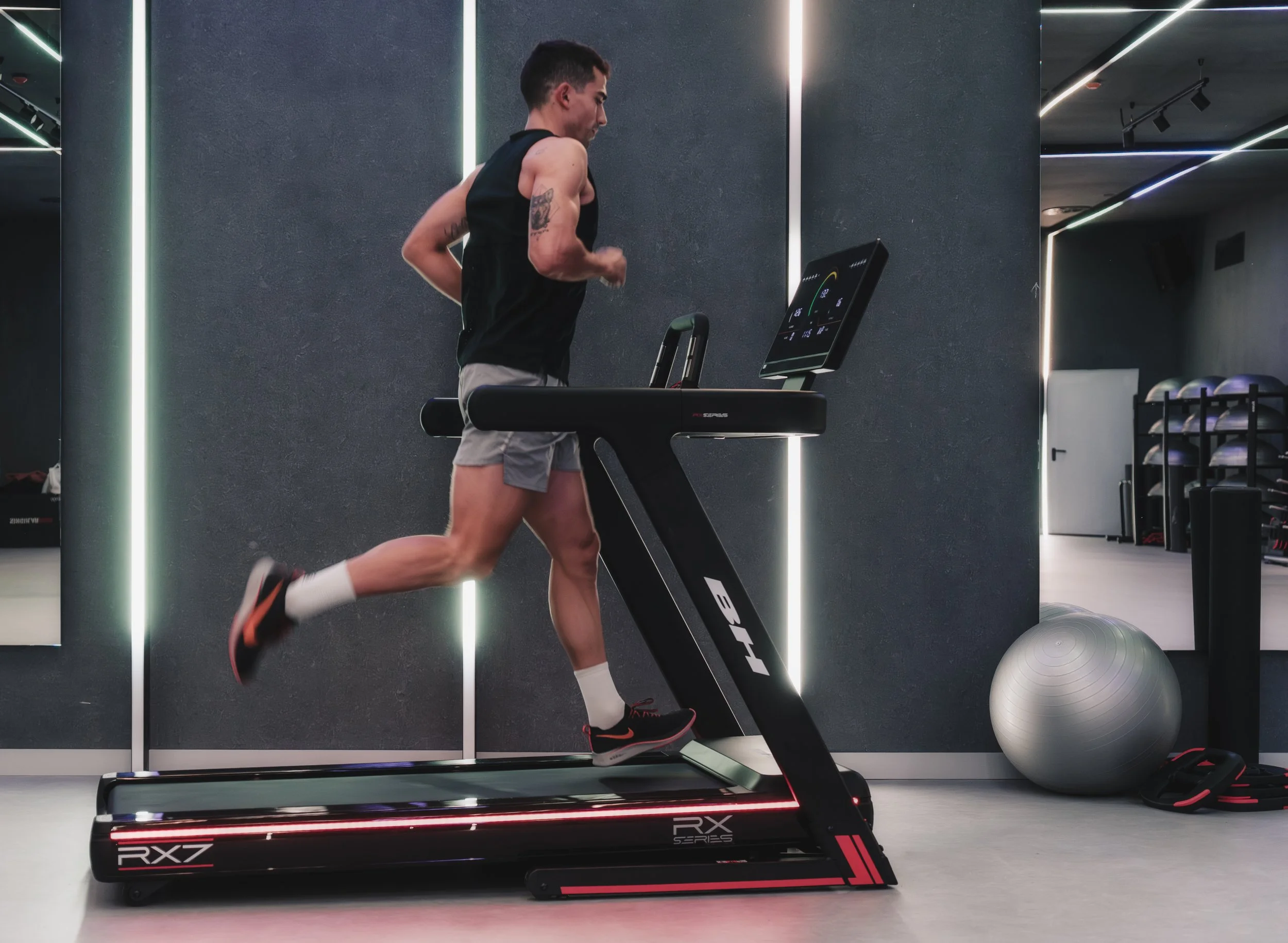 Man running on a treadmill in a modern gym, wearing black tank top and gray shorts.