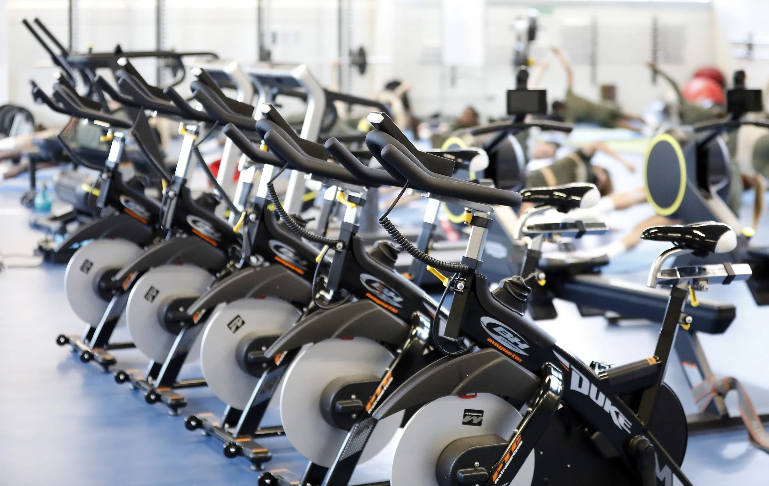 Indoor gym with multiple stationary exercise bikes in the foreground and people stretching on mats in the background.