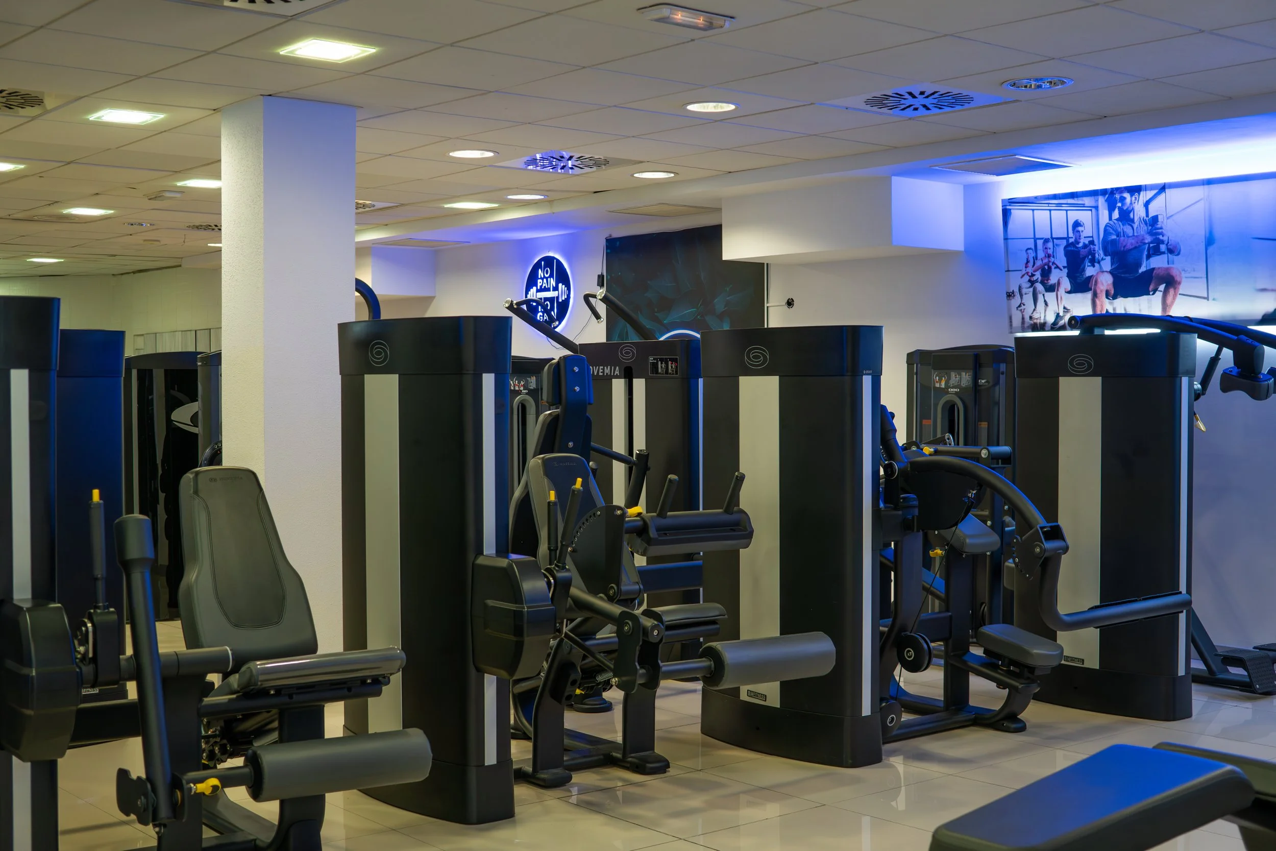 Fitness gym with various exercise machines in a well-lit room, some illuminated with blue lighting.