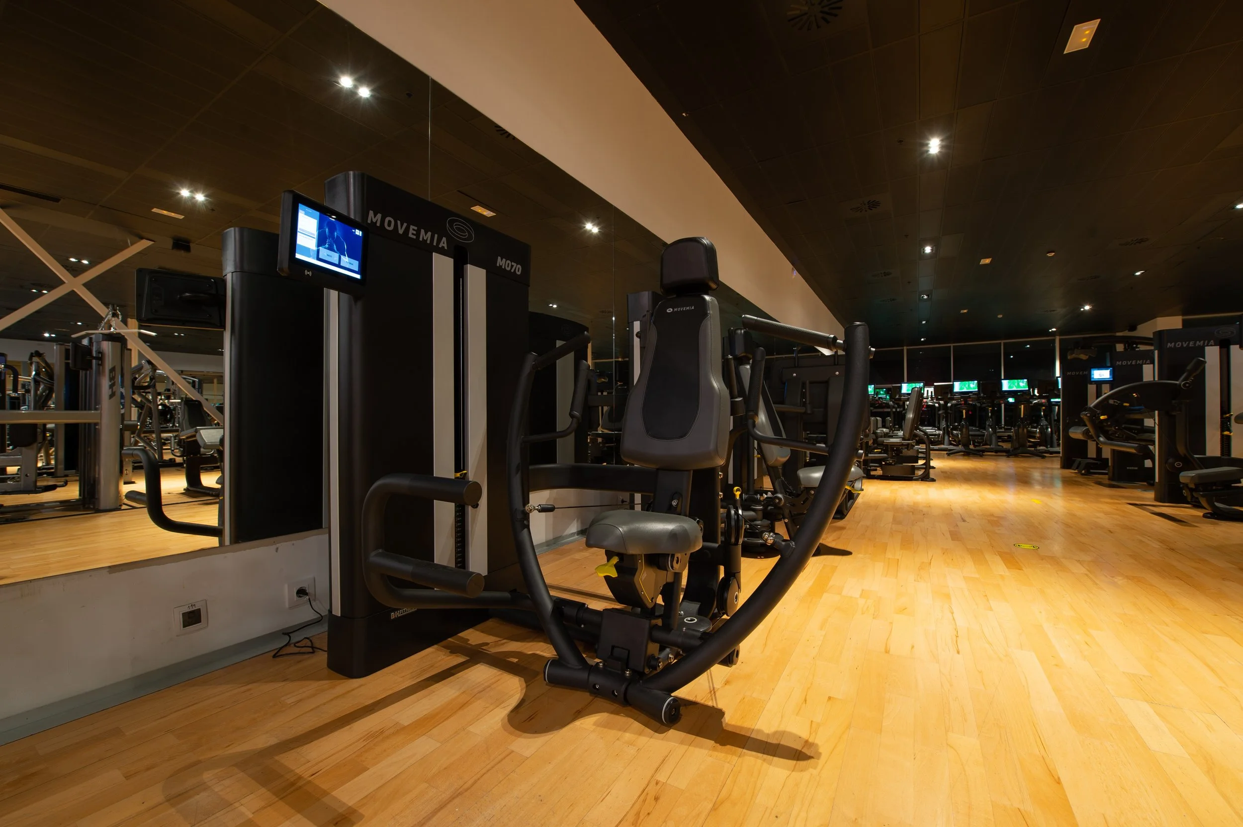 Empty gym with exercise machines and wooden flooring, featuring a large mirror and multiple TV screens in the background.