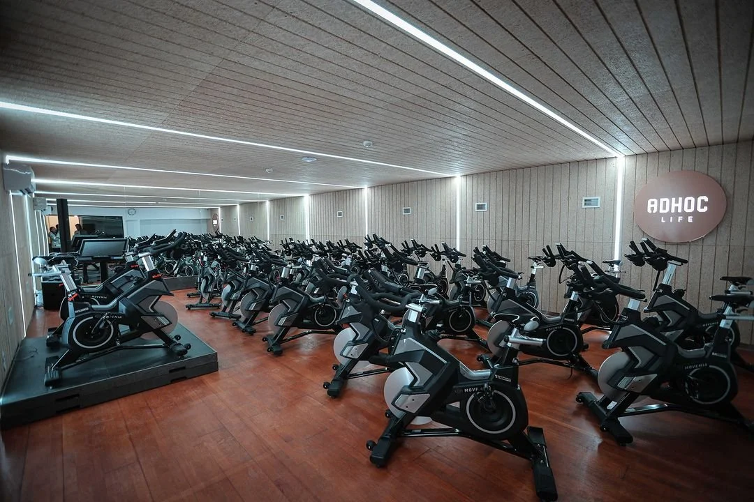 Indoor cycling studio with multiple stationary bikes arranged in rows, wooden ceiling, wall-mounted lights, and a digital display at the front, branded with 'ADHOC LIFE' sign on the wall.