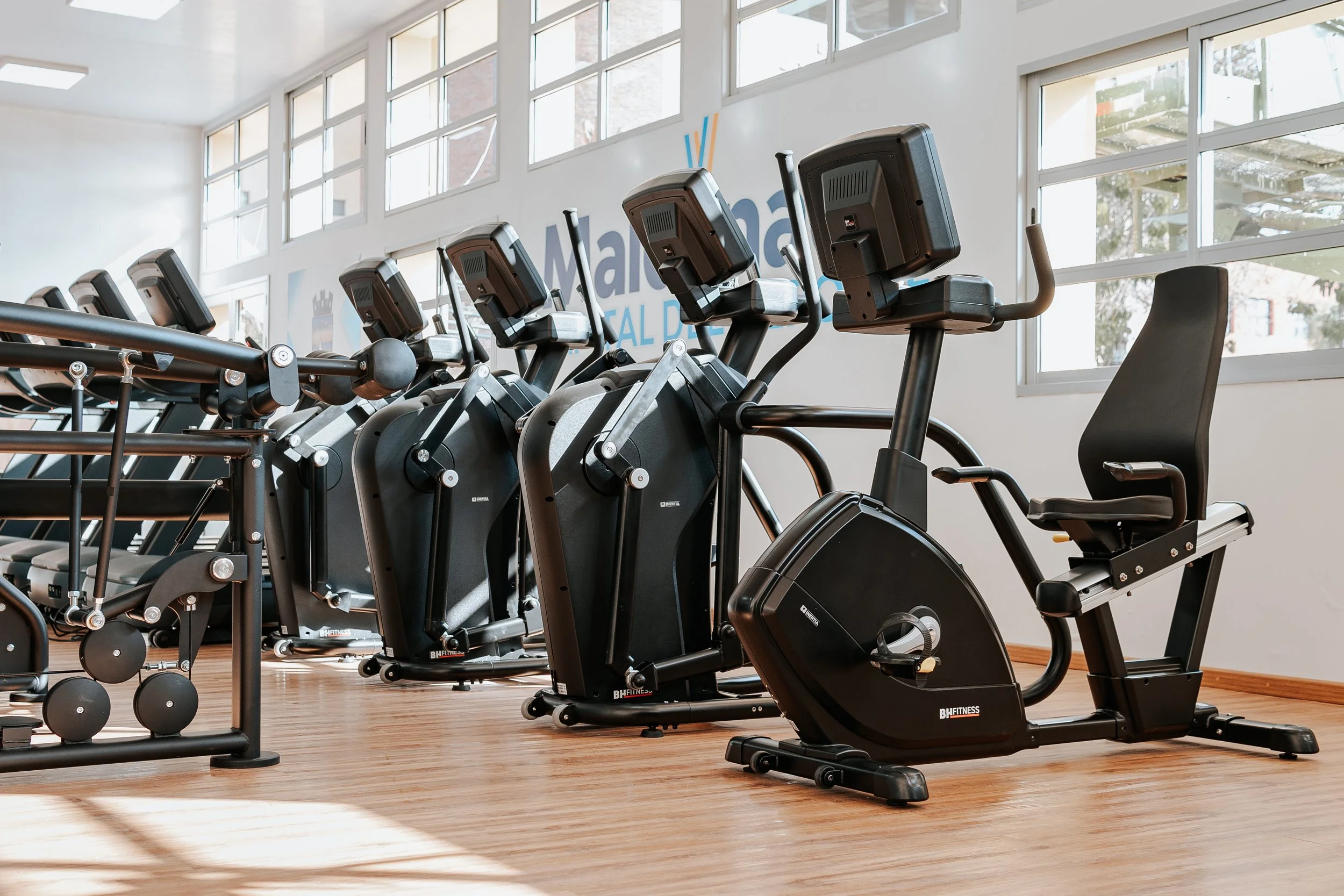 Row of exercise machines including stationary bikes in a gym with large windows.