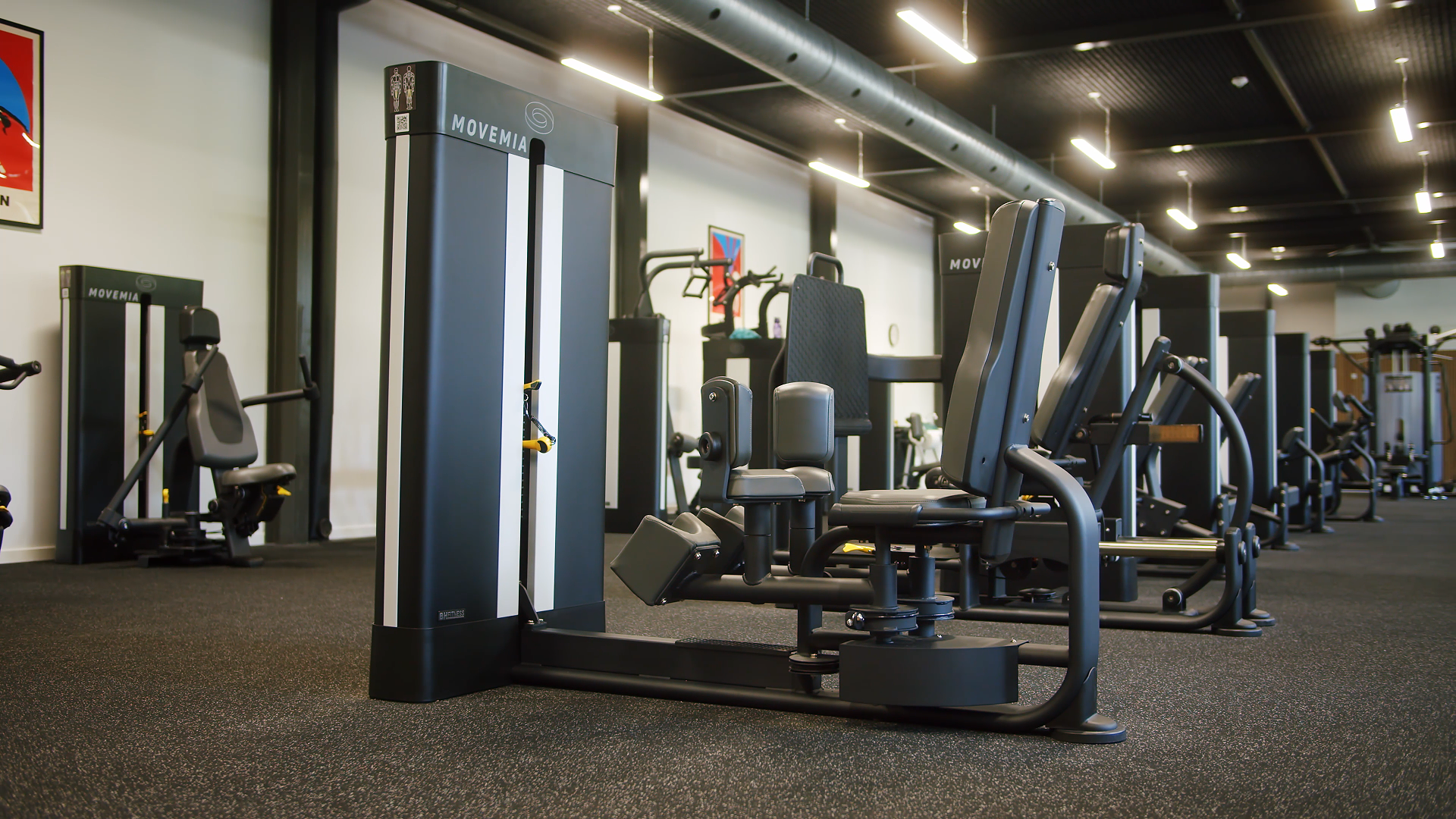 Empty gym with various exercise machines and equipment on a black carpeted floor, illuminated by ceiling lights.