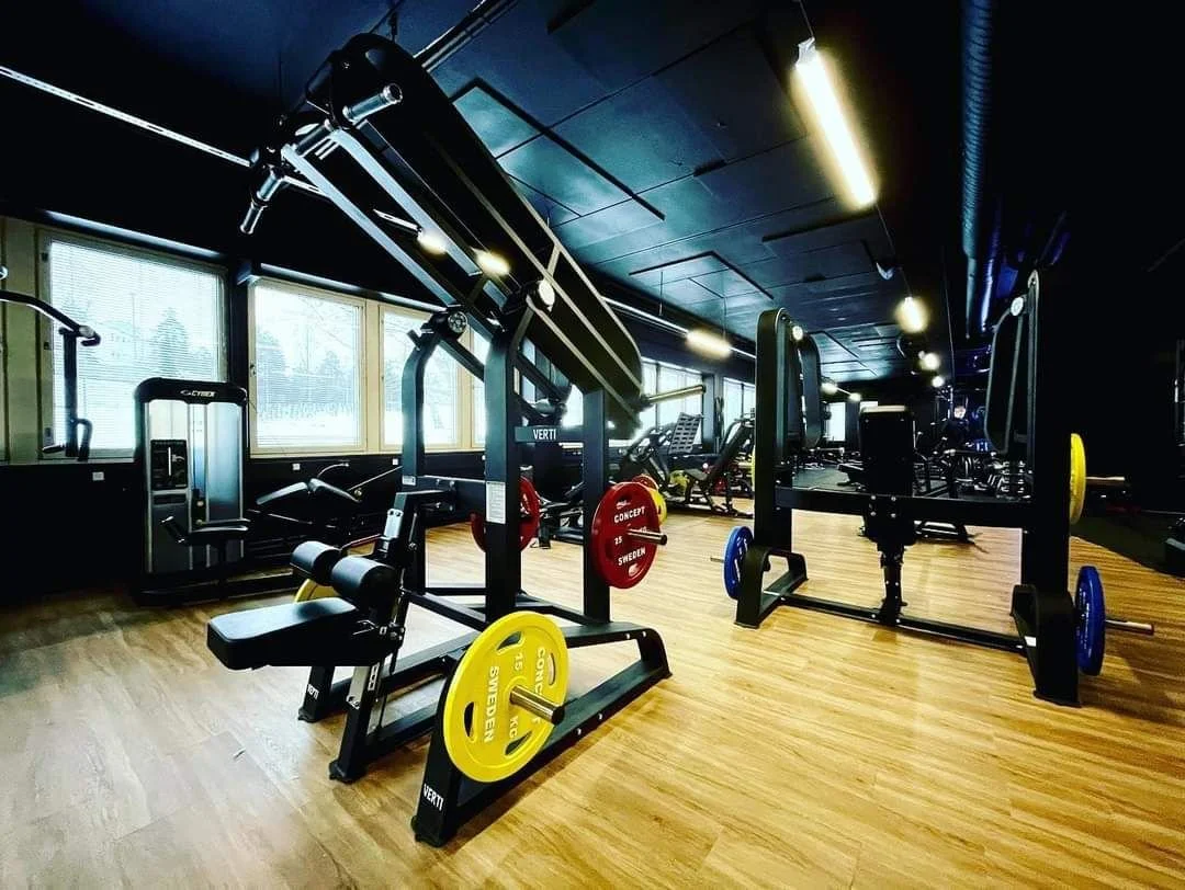 Gym with equipment including squat racks, weight plates, and cardio machines in a well-lit space with wooden floors and large windows.