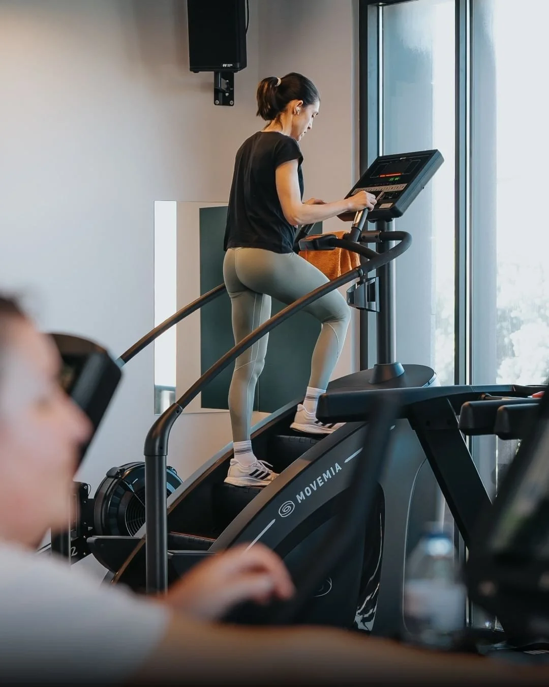 A woman running on a treadmill at a gym, wearing athletic clothes, with windows in the background.