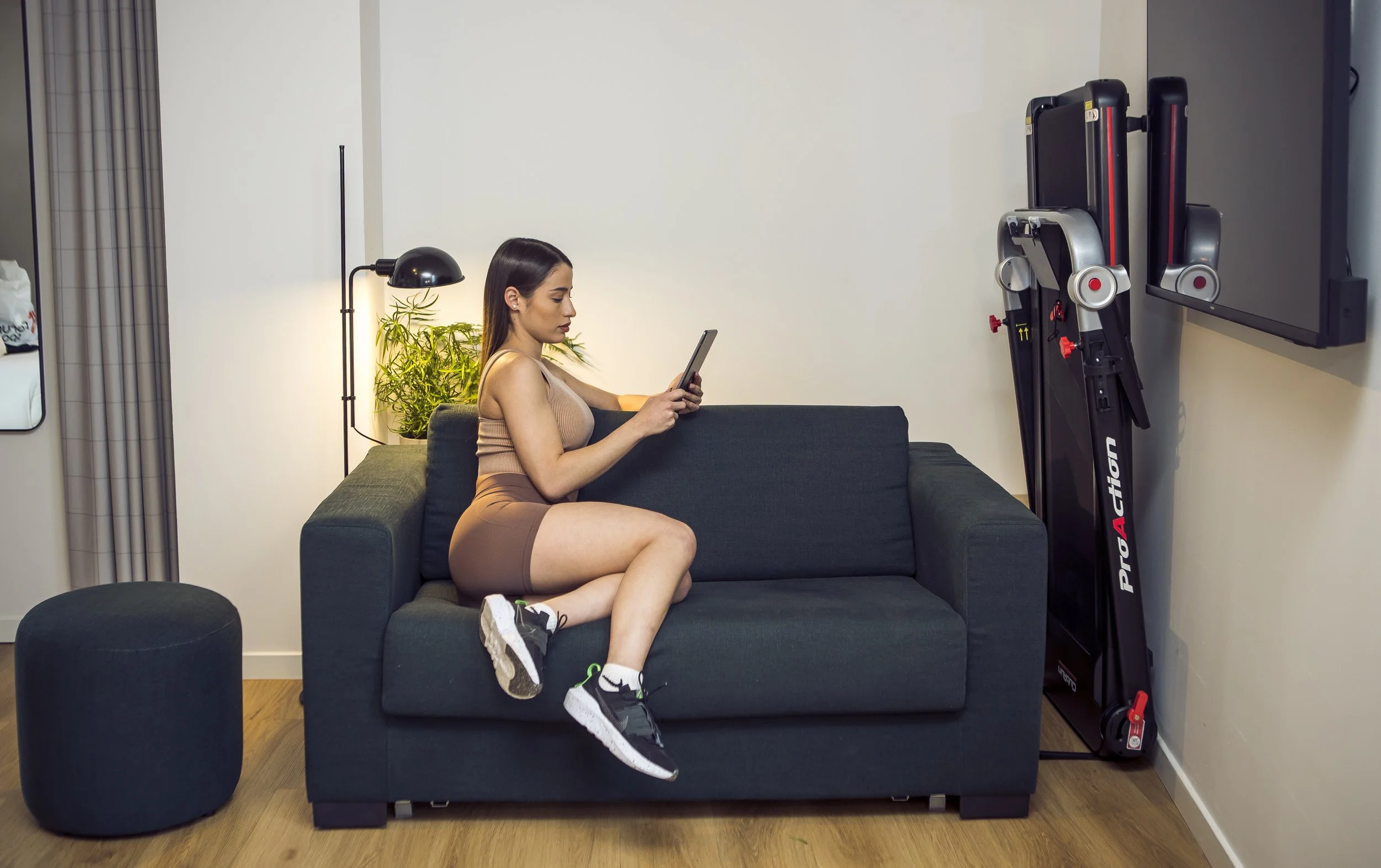 A woman sitting on a dark green sofa in a living room, looking at her phone. She is dressed in athletic wear and sneakers. There's a black ottoman nearby, a floor lamp with a plant behind it, and folded exercise equipment against the wall, with a TV