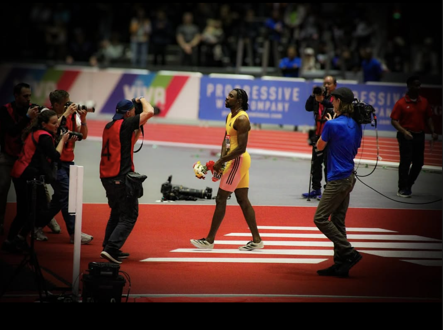 Athlete holding flowers walking across the finish line at a track event, surrounded by photographers and media personnel.