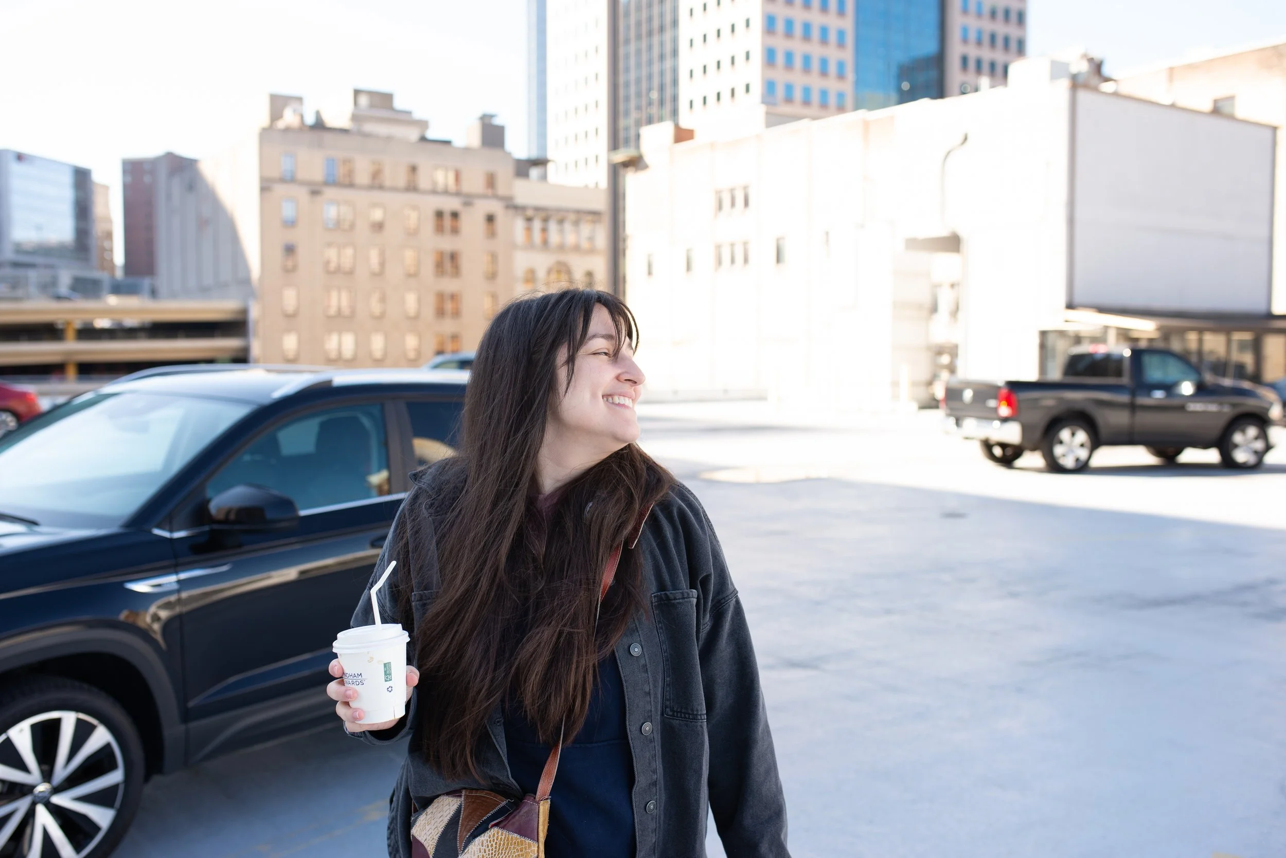 Person holding a coffee and smiling on a downtown Pittsburgh parking deck in early morning light, with city buildings in the background.