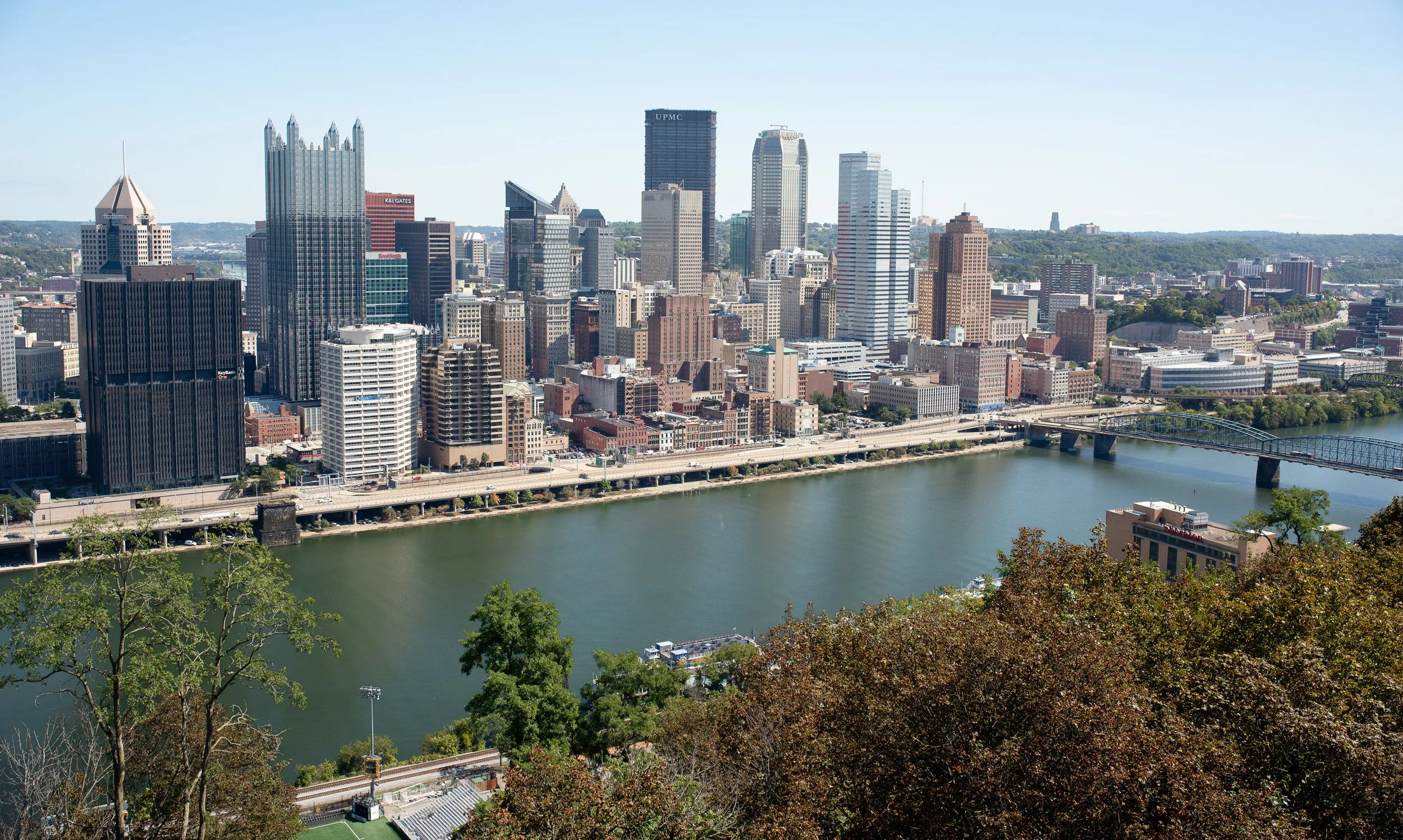Panoramic view of the Pittsburgh skyline from Grandview Overlook, with downtown buildings, bridges, and the river framed by trees in the foreground.