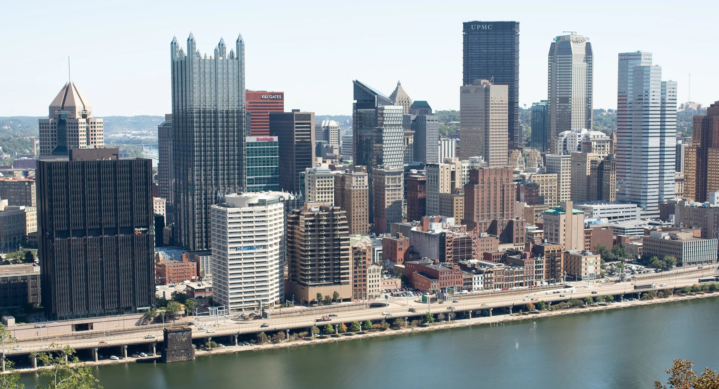 Close view of downtown Pittsburgh skyline along the river, with high-rise buildings and bridges visible from an elevated overlook.