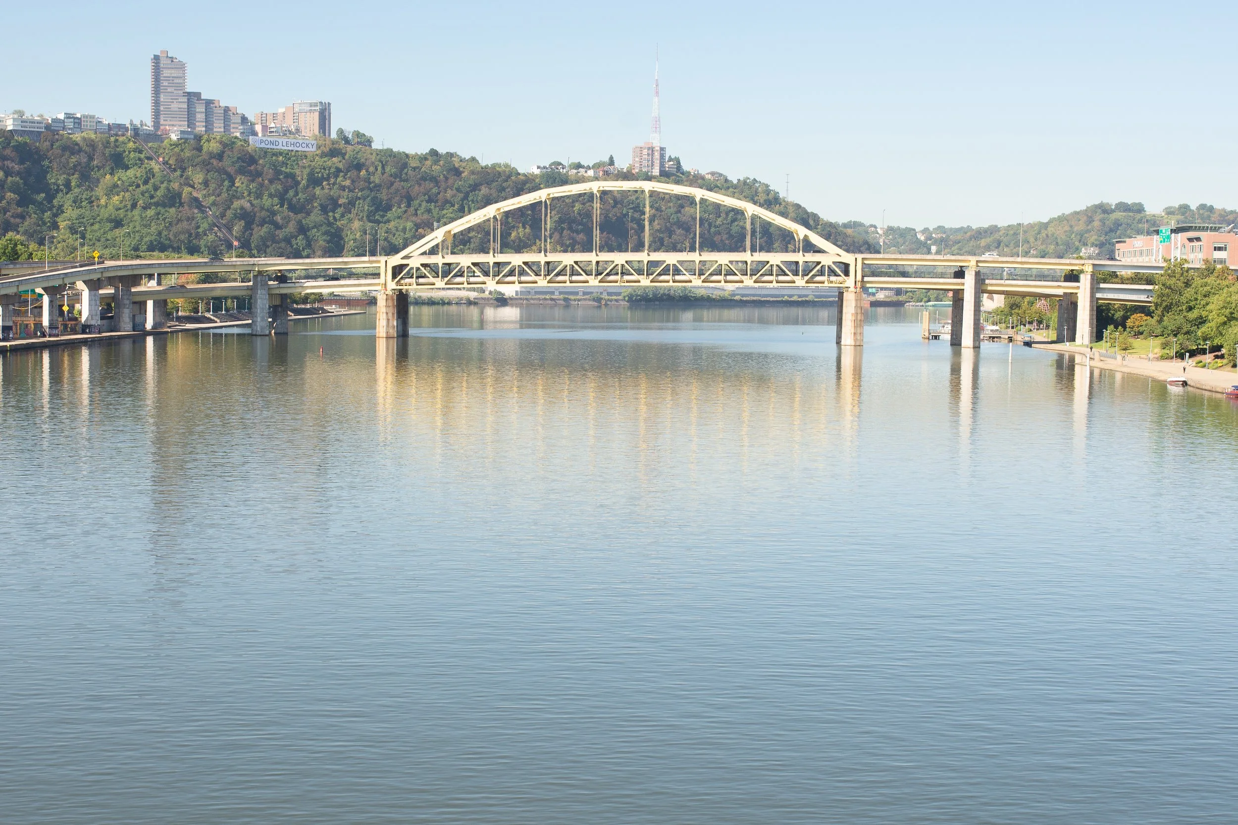 View of the West End Bridge spanning a calm river, with tree-covered hills and city buildings in the distance under clear skies.